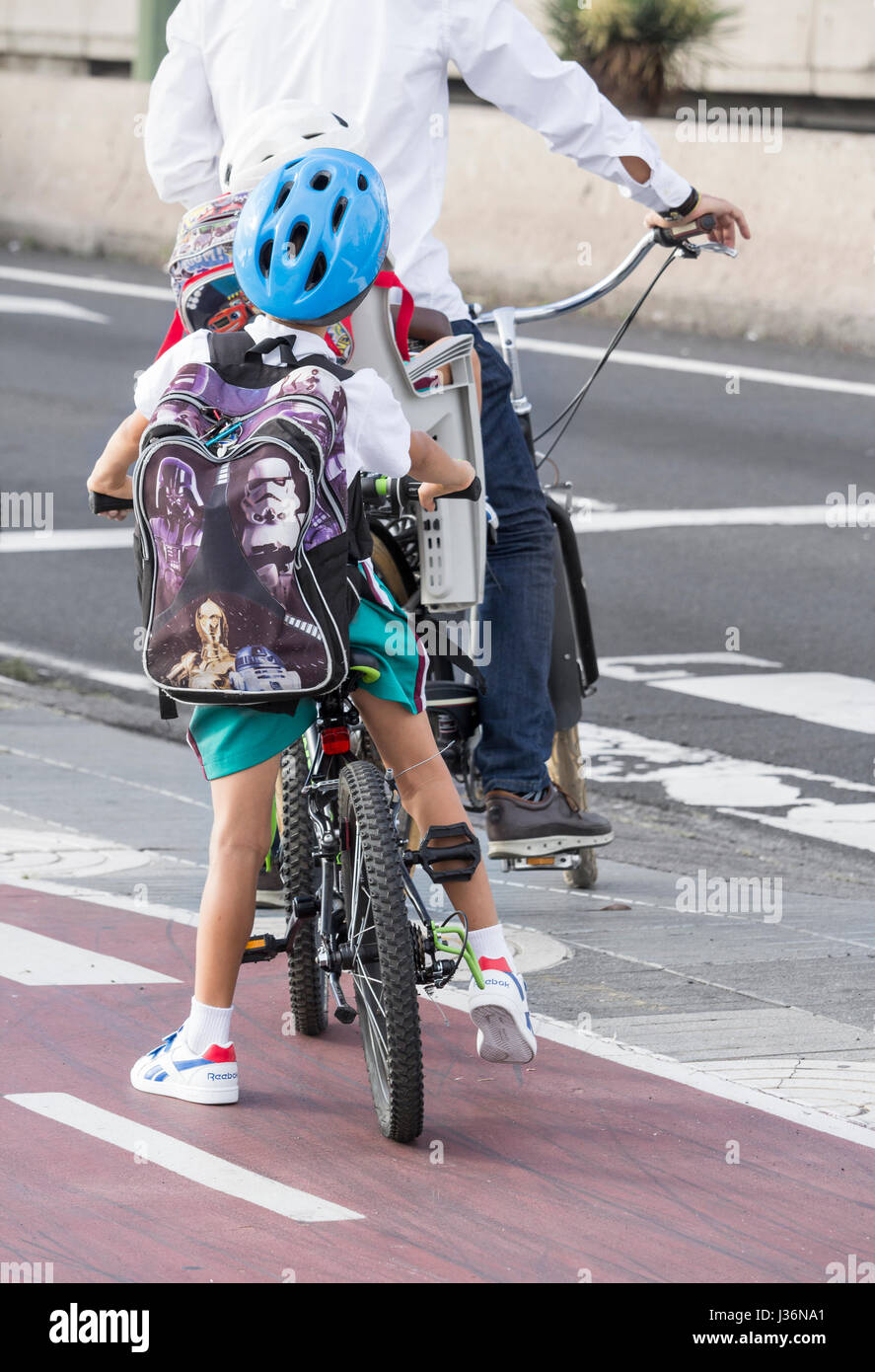 Young Spanish boy in school uniform riding to school on bicycle behind adult who is carrying Stock Photo