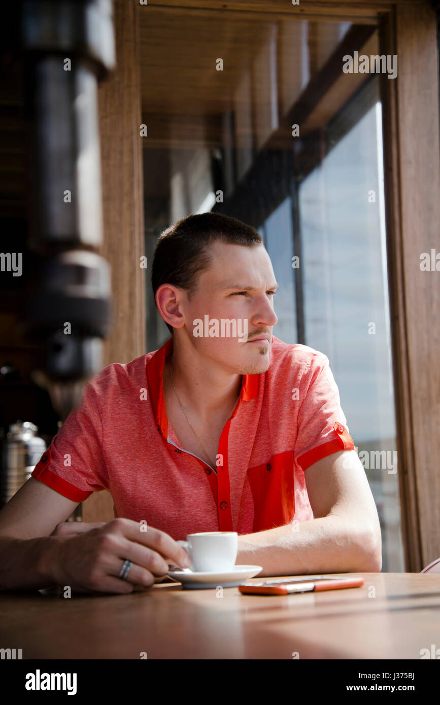 A sensitive young man with red shirt looking thoughtfully out of the window while sitting at a table with a cup of coffee in hand, reflecting on the f Stock Photo