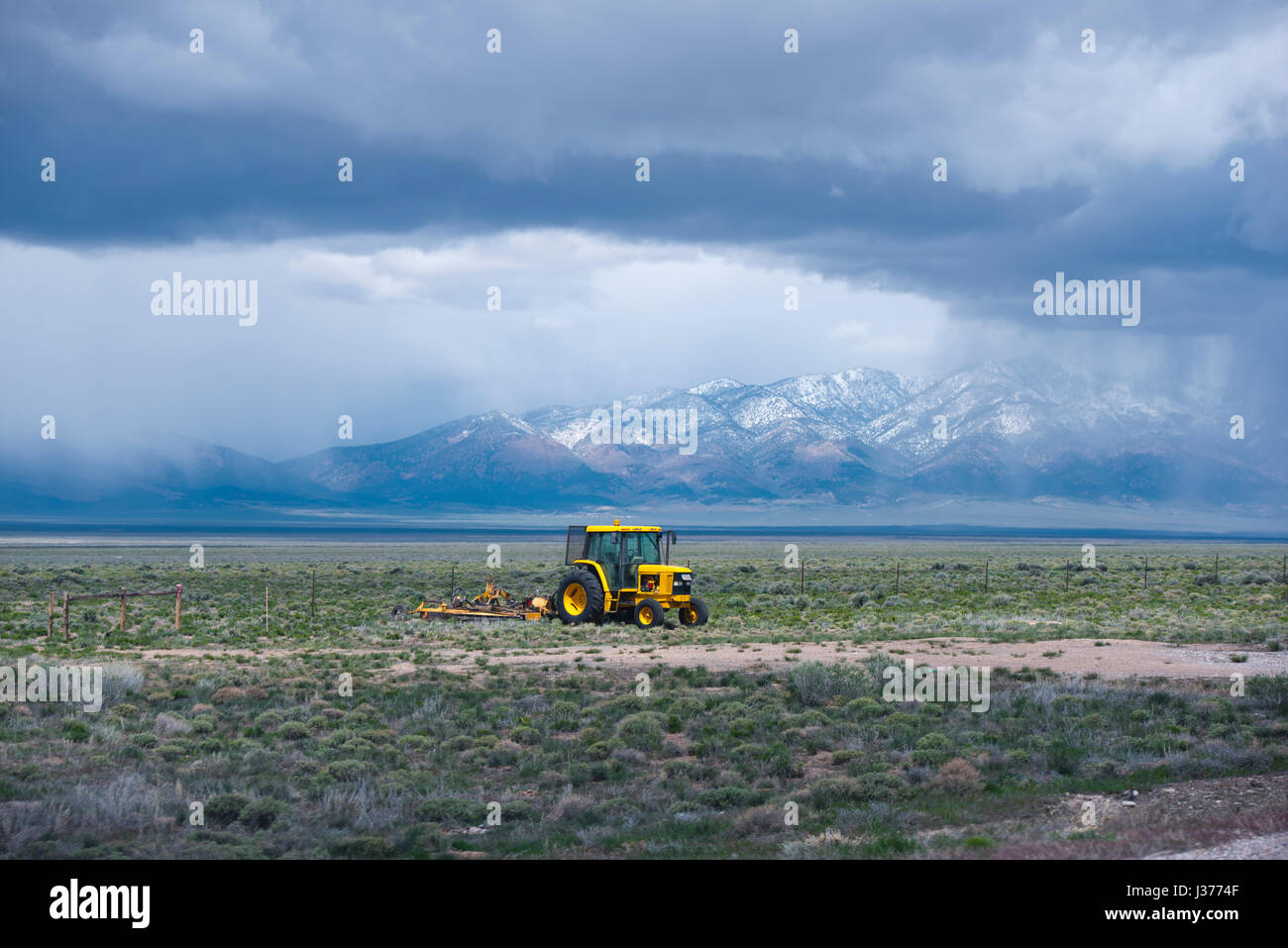Yellow tractor with a plow working in the field in the middle of Nevada, cultivating land for growing food plants, framed by a ridge of high mountains Stock Photo