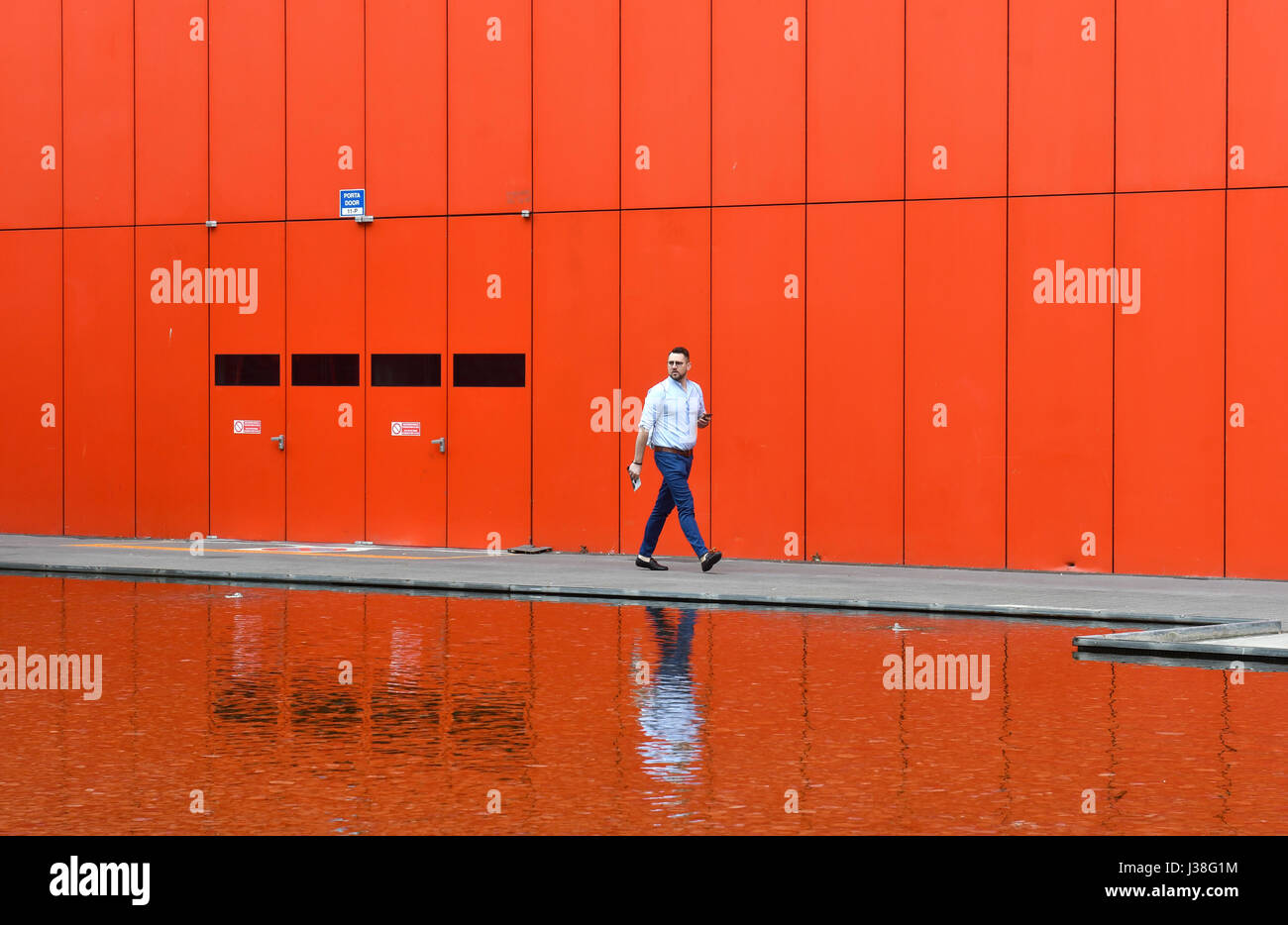 Man walking with red building in the backgroung reflected on a water at Fiera Rho Pero, in Milan. Stock Photo