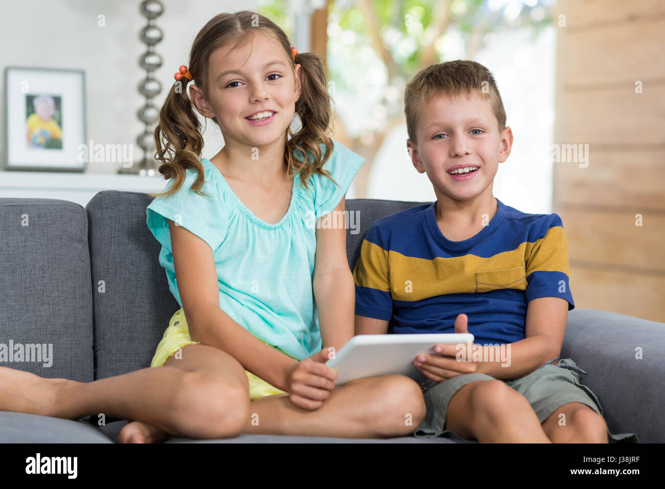 Portrait of siblings with digital tablet sitting on sofa in living room ...