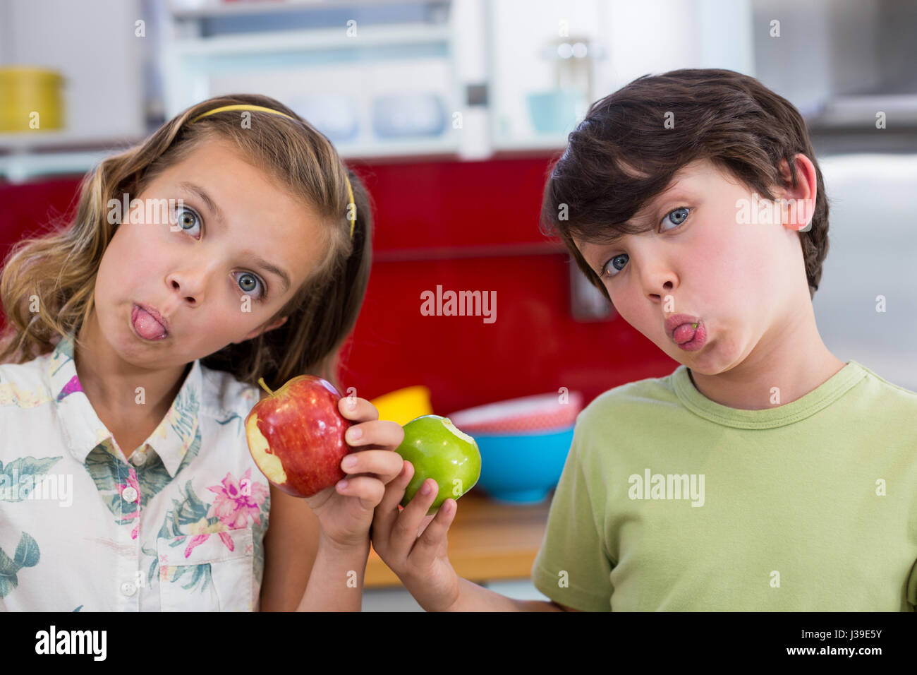 Siblings holding apple and pulling funny faces in kitchen at home Stock Photo