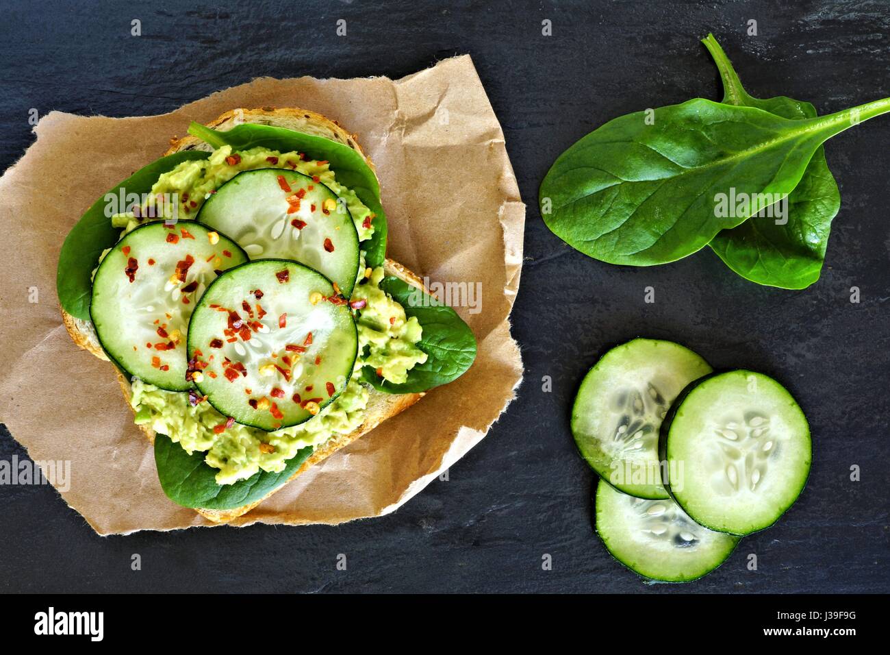 Avocado toast with cucumber, spinach and whole grain bread on paper against a dark slate background Stock Photo