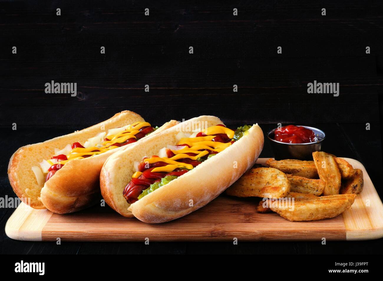 Two hot dogs fully loaded with toppings and potato wedges on wooden board with dark background Stock Photo
