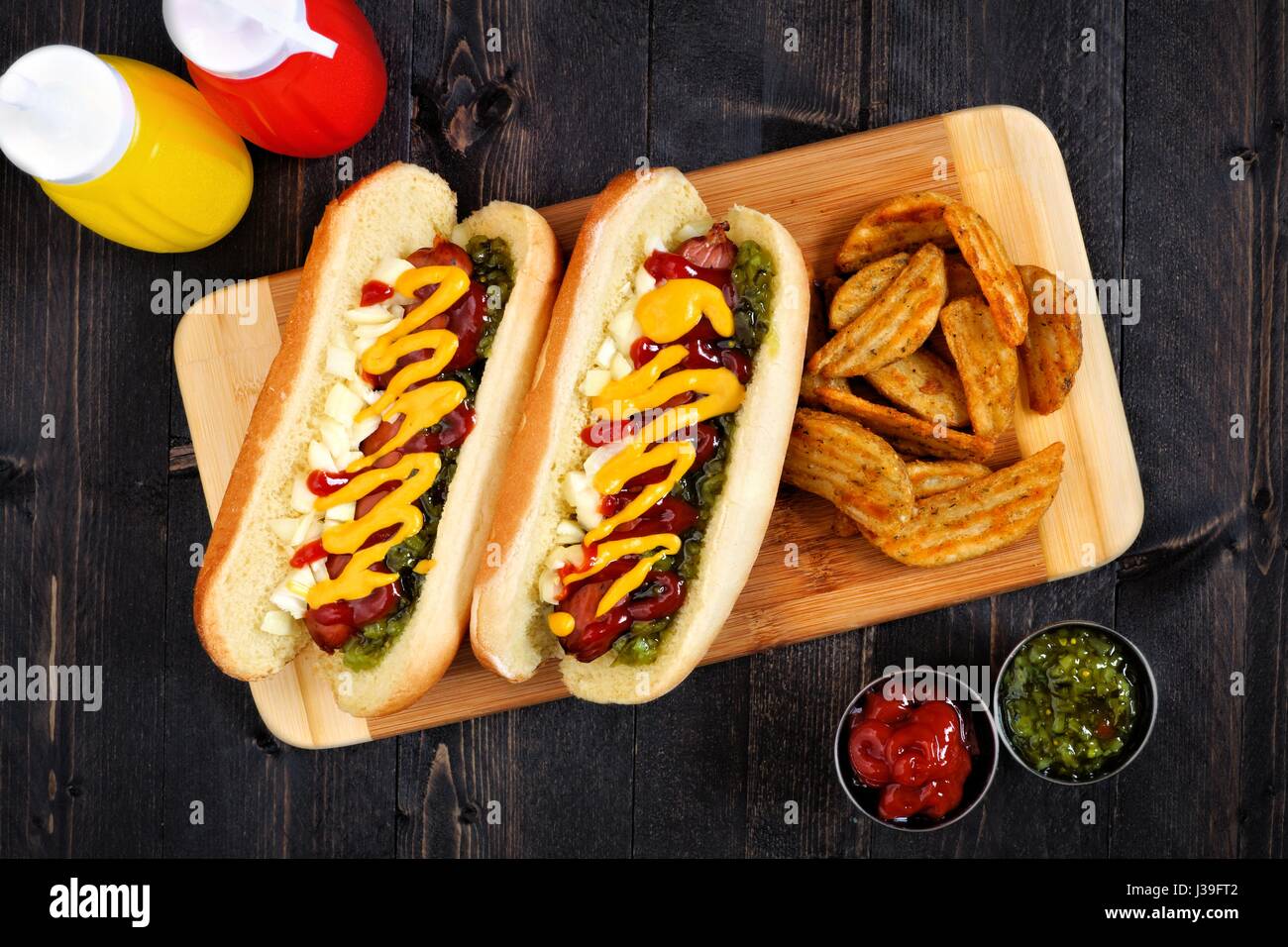 Two hot dogs fully loaded with toppings and potato wedges on wooden board, overhead scene Stock Photo