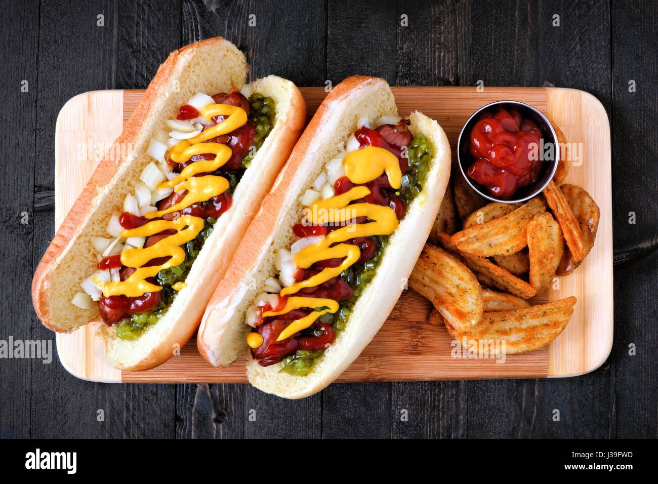 Two hot dogs fully loaded with toppings and potato wedges on wooden board, overhead view Stock Photo