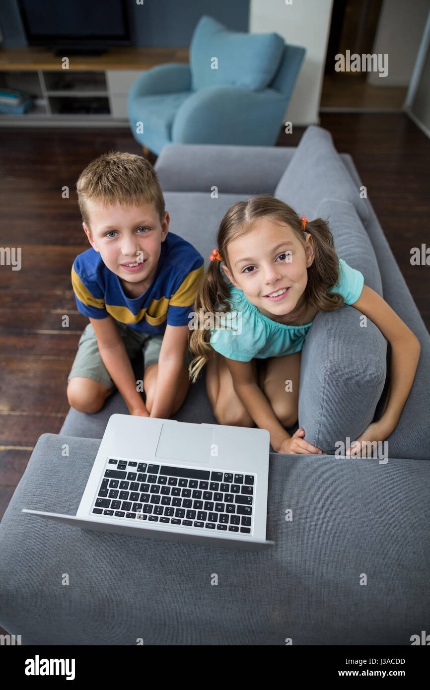 Portrait of siblings with laptop sitting on sofa in living room at home ...