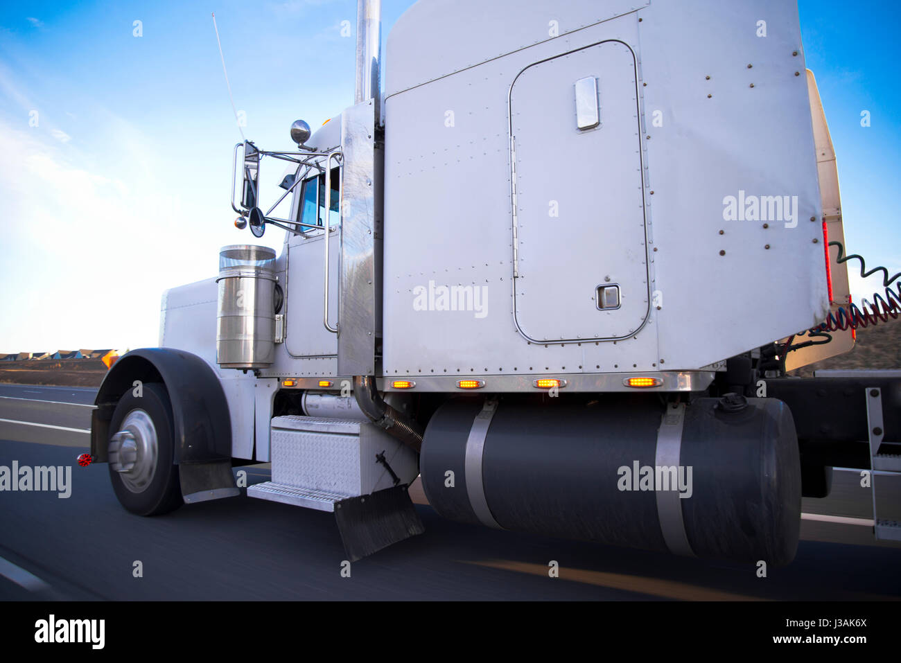 A classic huge, powerful white truck for long distance haulage with a long cab and huge fuel tank is speeding to its destination on the straight road Stock Photo