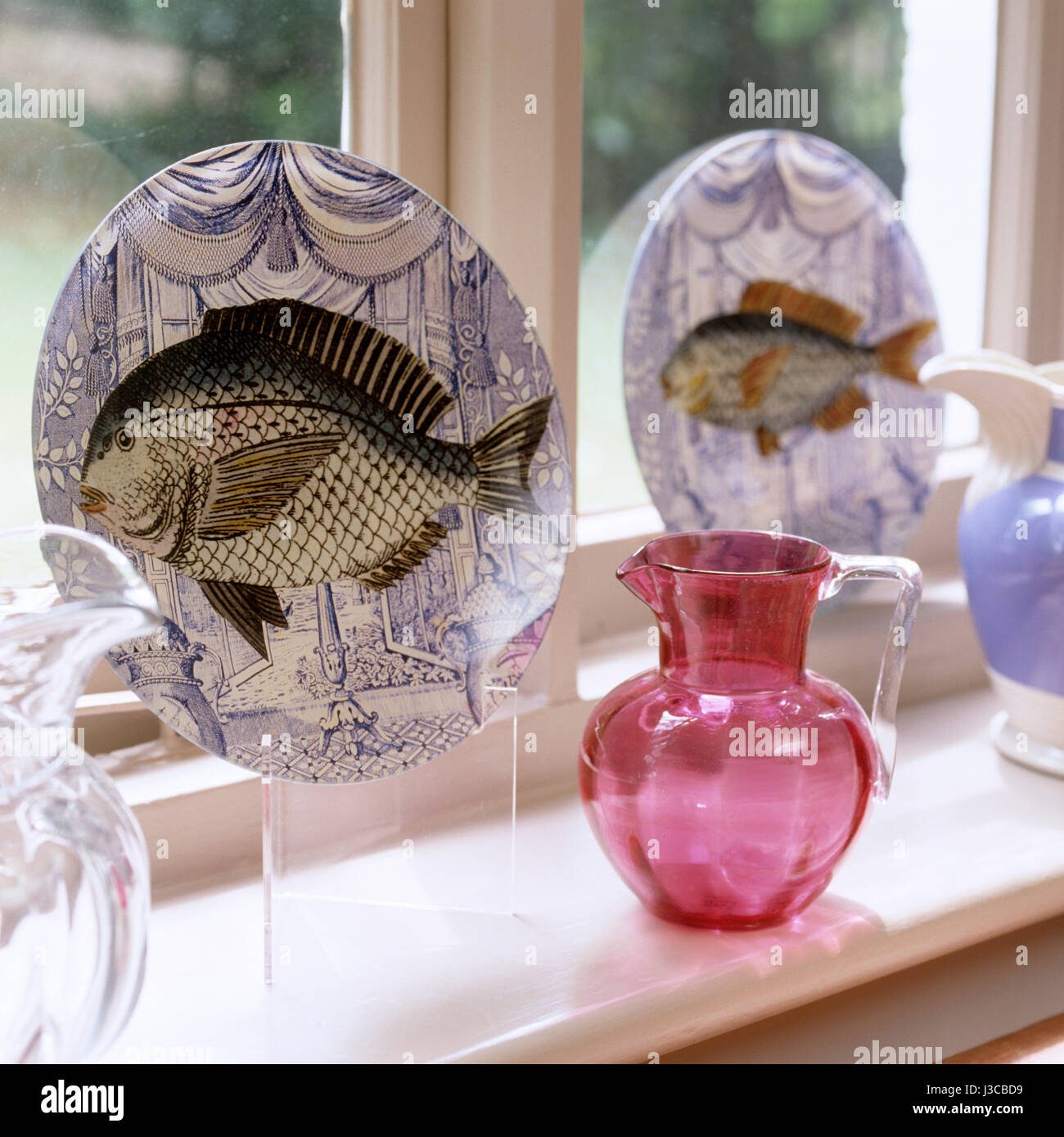 Ornamental plates with fish illustrations on window sill with pink glass jug Stock Photo