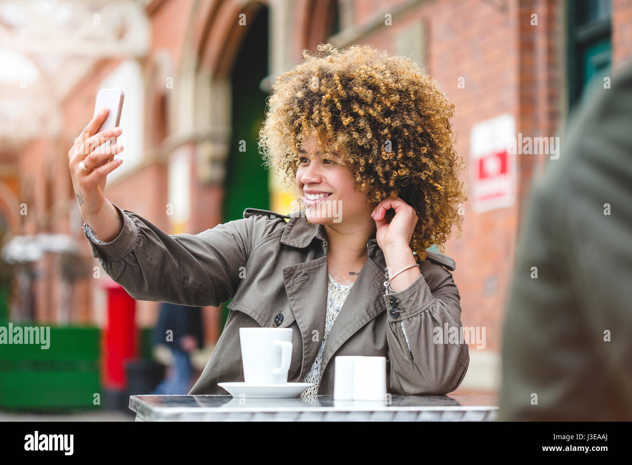 Woman taking a selfie on a smartphone. She is sat outside a cafe and is using a smart phone to take a photo. Stock Photo