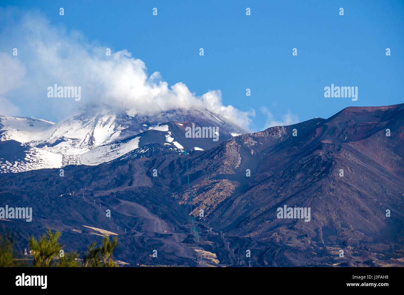 Mount Etna, an active stratovolcano, from the south with the smoking peak, snowy flanks, a lateral crater in the centre and a cable car line in the Pr Stock Photo