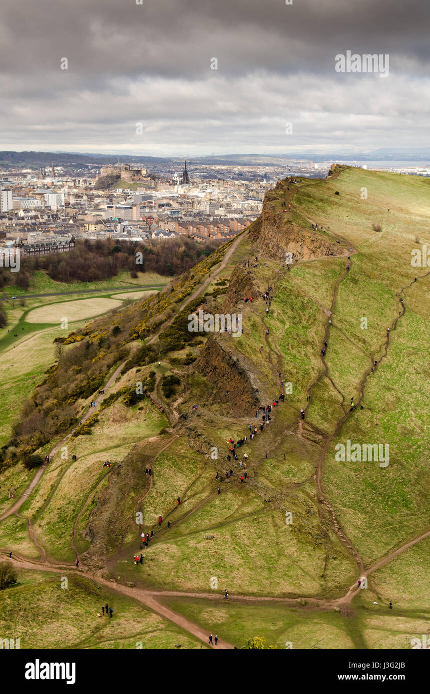 A crowd of people walk on Salisbury Crags, rising spectacularly in Holyrood Park from the cityscape of Edinburgh's Old Town and castle. Stock Photo