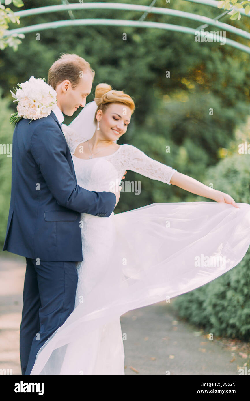 Happy young gentle groom and bride posing at the park. Bridal wedding bouquet of flowers in hands Stock Photo