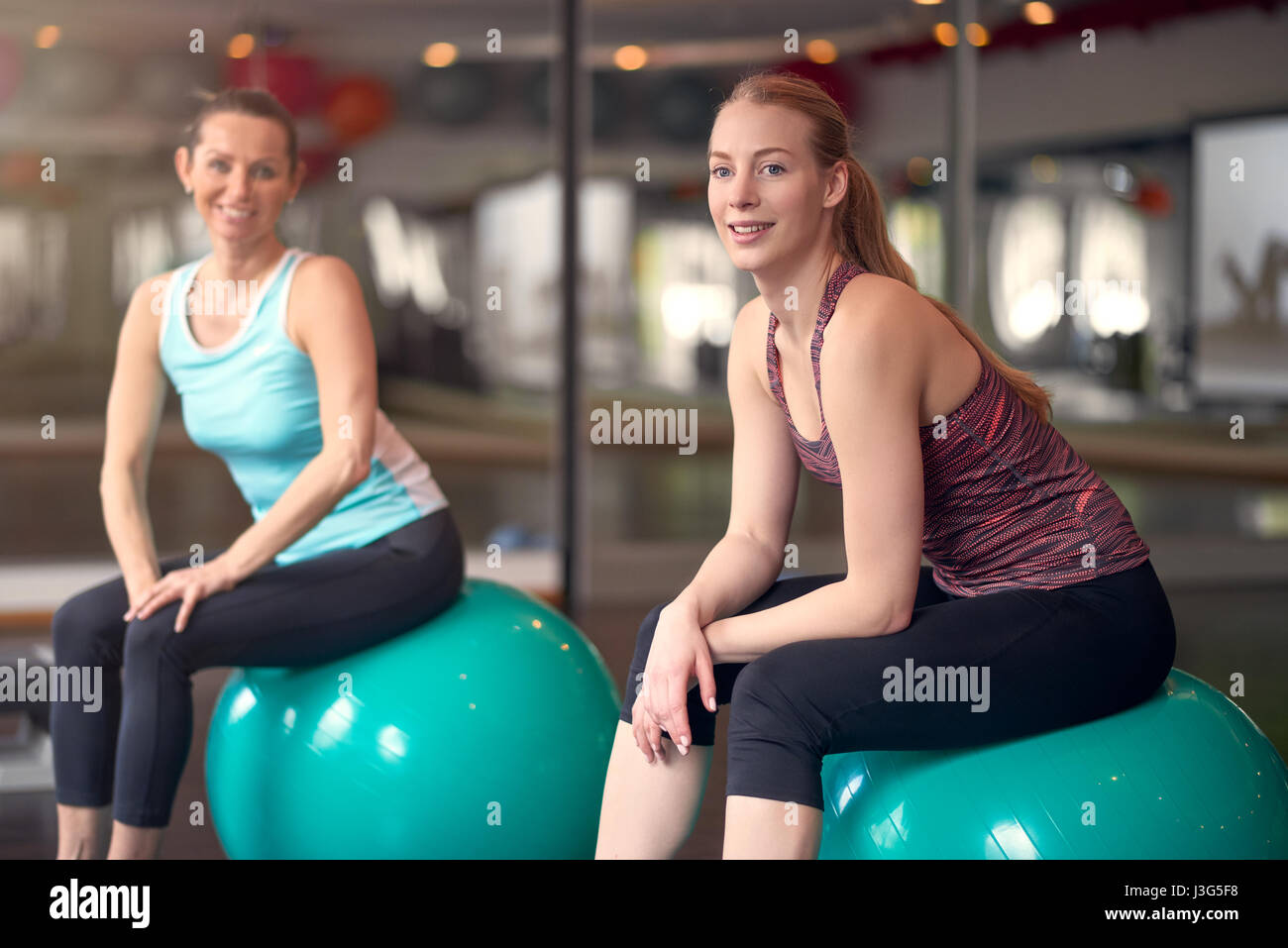 Two healthy young women with pilates or gym balls taking a break from their workout in a gym sitting smiling at the camera in an active lifestyle and  Stock Photo