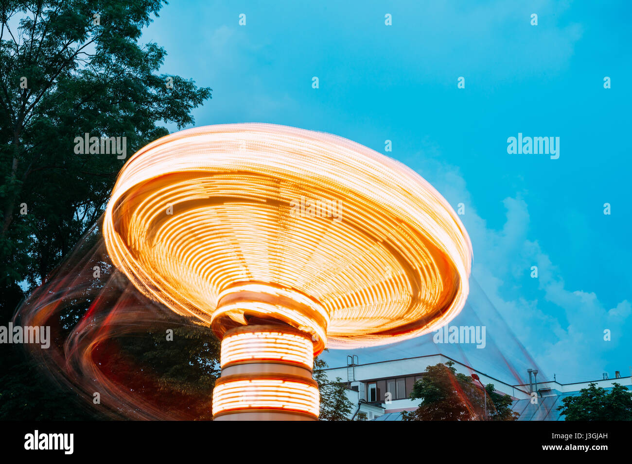 Top Disk Part Of Brightly Illuminated, Dynamic Energy Rotating High Speed Carousel Merry-Go-Round With Blurred Motion Effect Around. Blue Evening Sky  Stock Photo
