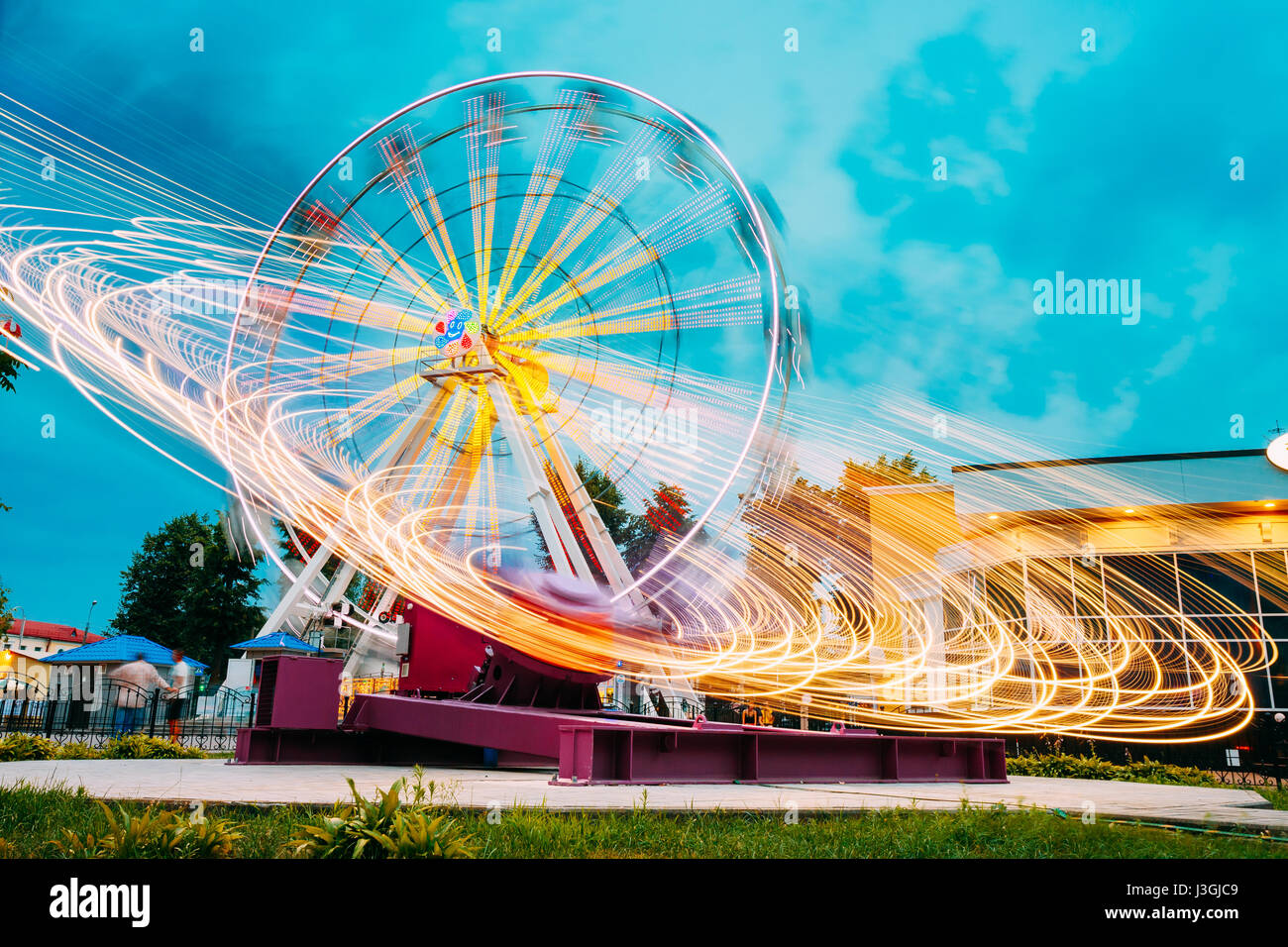 Motion Blurred Effect Around Of High Speed Rotating Illuminated Attraction Feature In City Amusement Park. Ferris Wheel On Summer Evening Blue Sky Bac Stock Photo