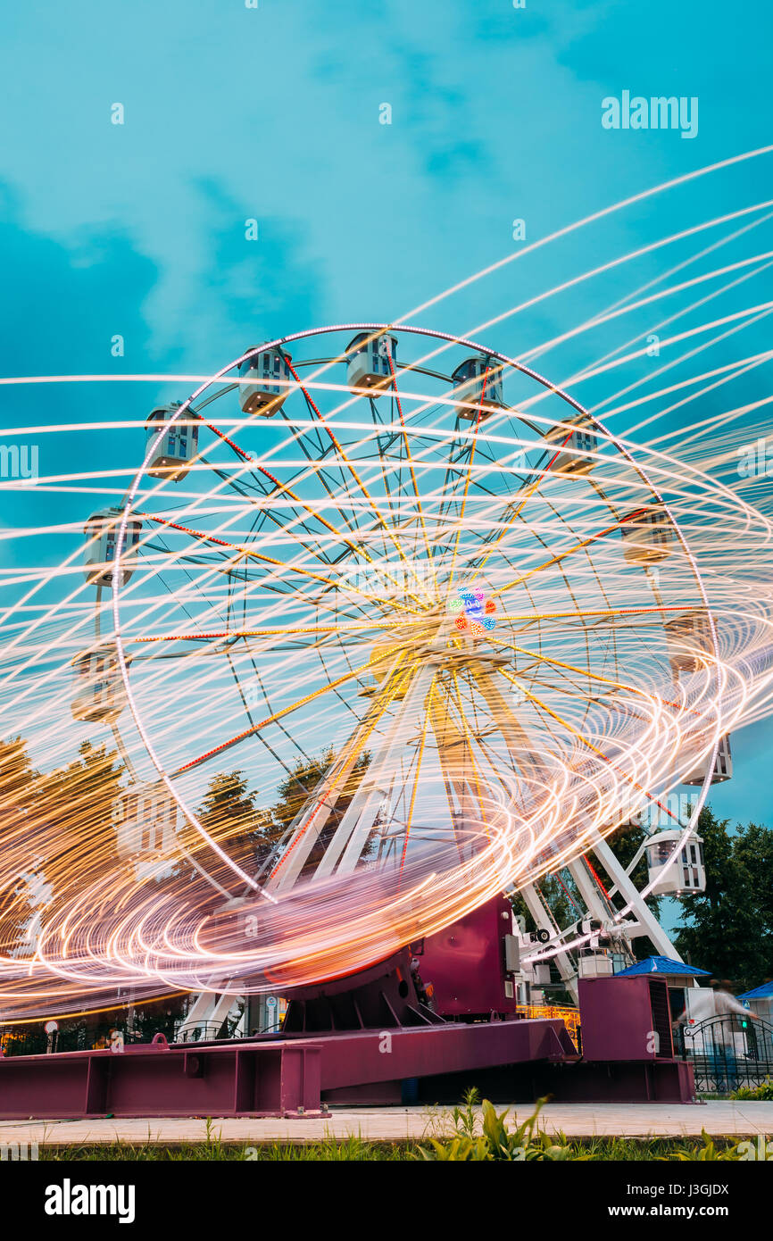 Motion Blurred Effect Around Of High Speed Rotating Illuminated Attraction Feature In City Amusement Park. Ferris Wheel On Summer Evening Blue Sky Bac Stock Photo