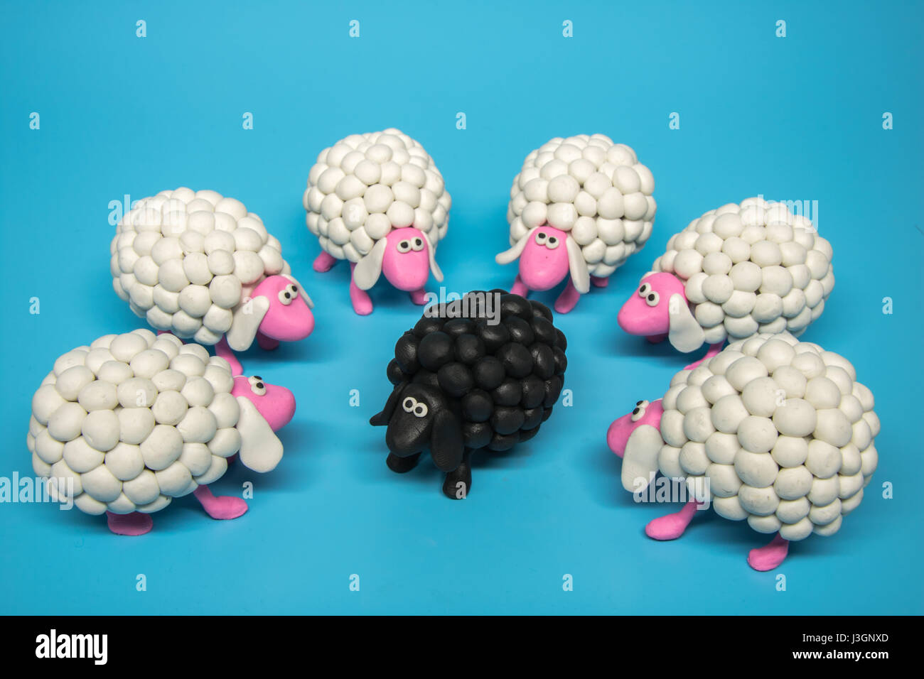 Concept - A circle of white sheep surrounds one black sheep, on a solid blue background. Stock Photo