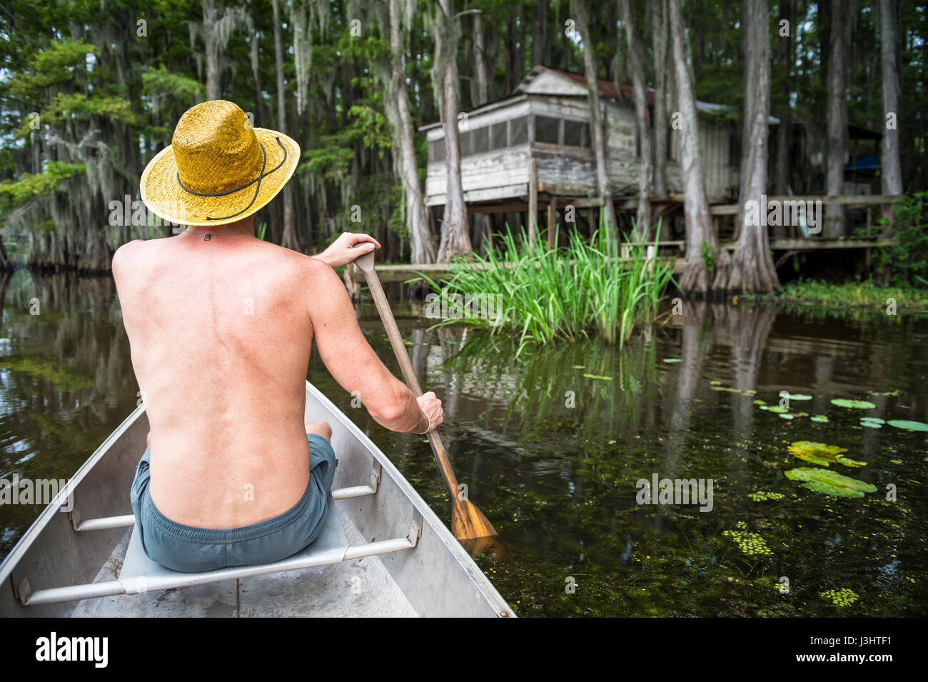 Man with hat sitting at the front of a canoe paddling into a dark swamp scene in Caddo Lake, Texas Stock Photo