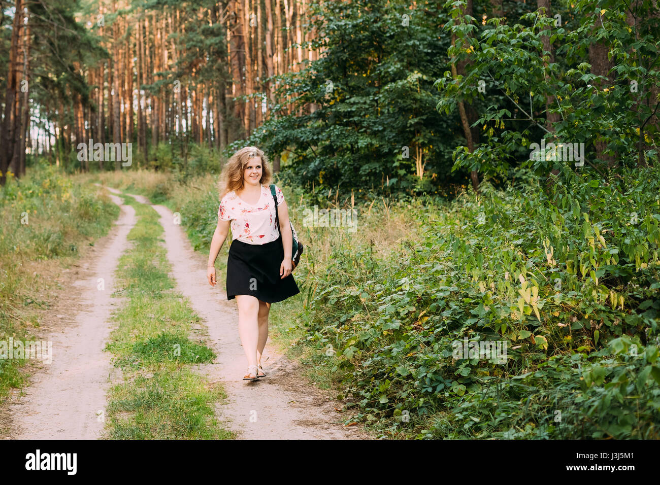 Young Pretty Plus Size Caucasian Happy Smiling Girl Woman In White T-Shirt And Black Short Skirt Walking Full-Length On Road In Summer Pine Forest Stock Photo