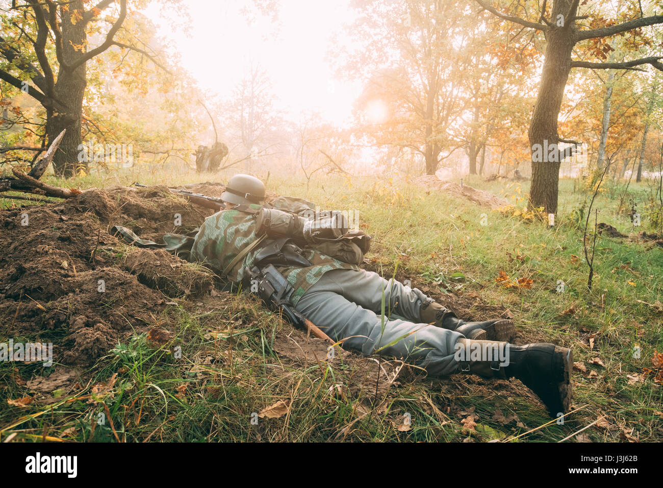 Unidentified Re-enactor Dressed As German Wehrmacht Infantry Soldier In World War II Hidden Sitting With Rifle Weapon In An Ambush In Trench In Autumn Stock Photo