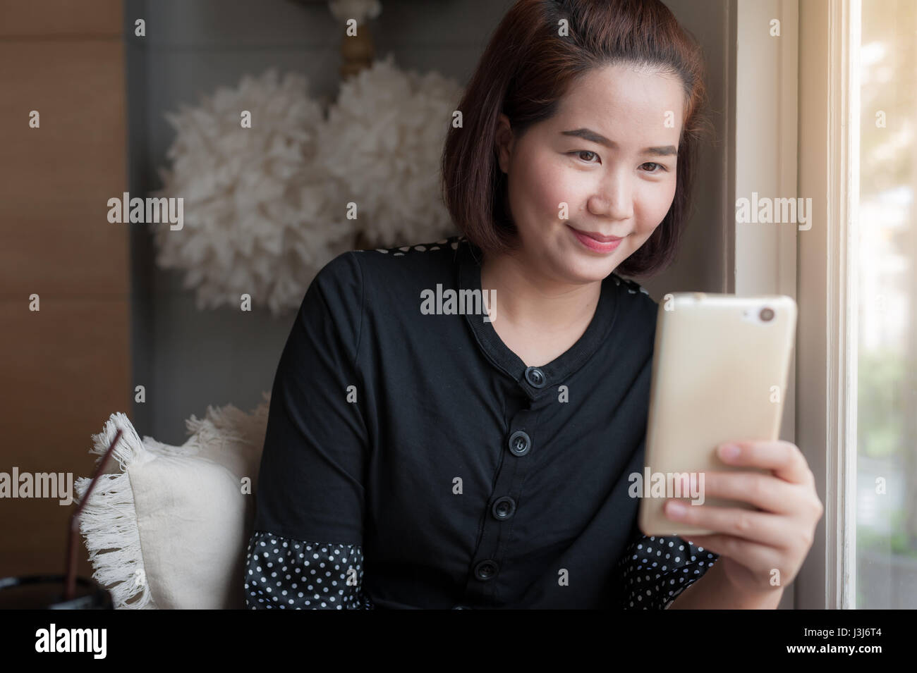 Young Asian woman use smartphone for selfie beside window in cafe with good mood. Trendy lifestyle with technology and internet Stock Photo