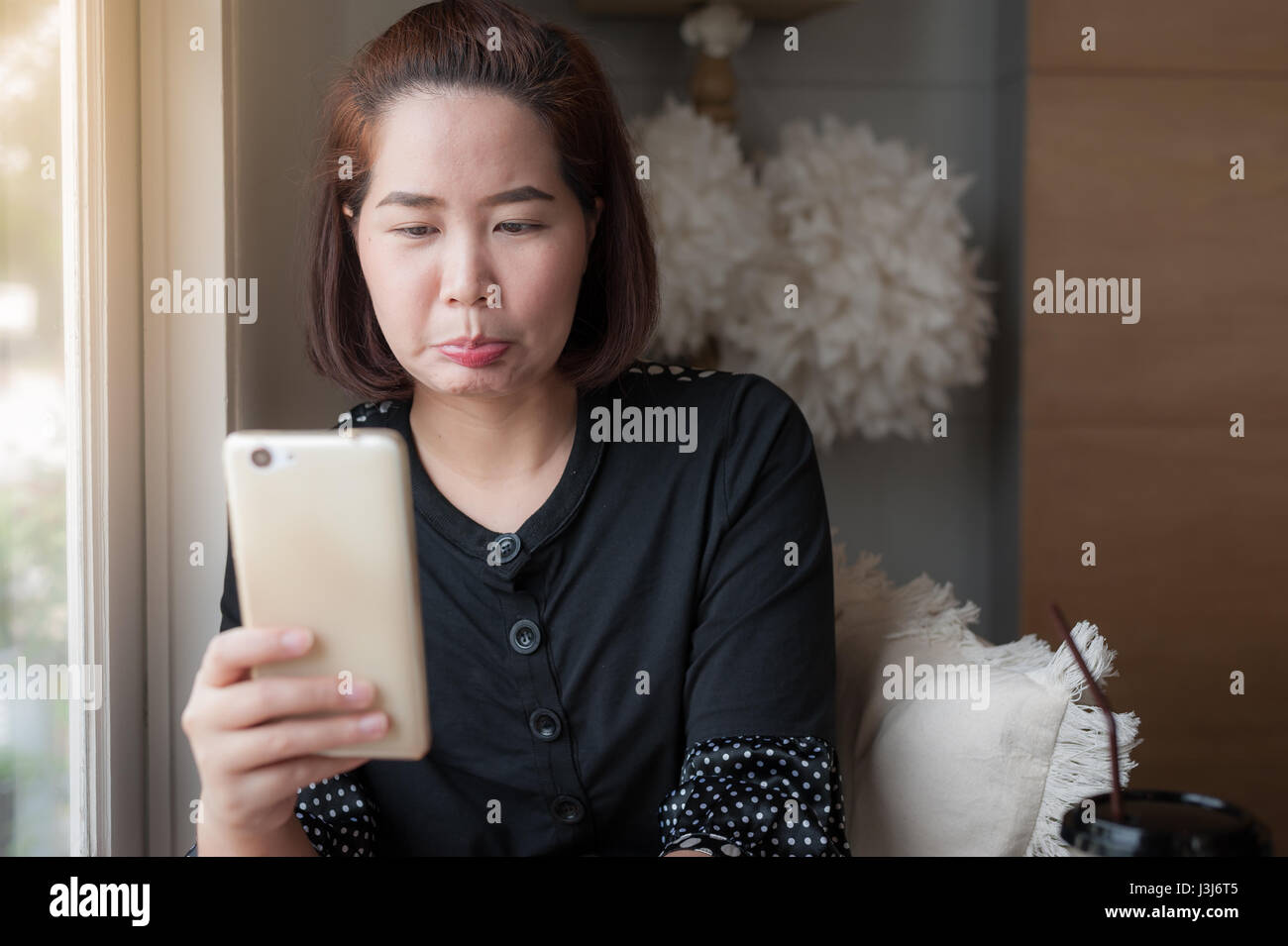 Young Asian woman use smartphone for selfie beside window in cafe with good mood. Trendy lifestyle with technology and internet Stock Photo