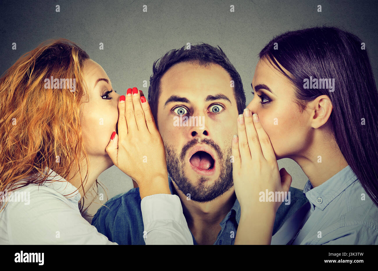 Two young women telling whispering secret gossip in the ear to an amazed shocked man with wide open mouth. Human emotions face expression reaction. Ho Stock Photo