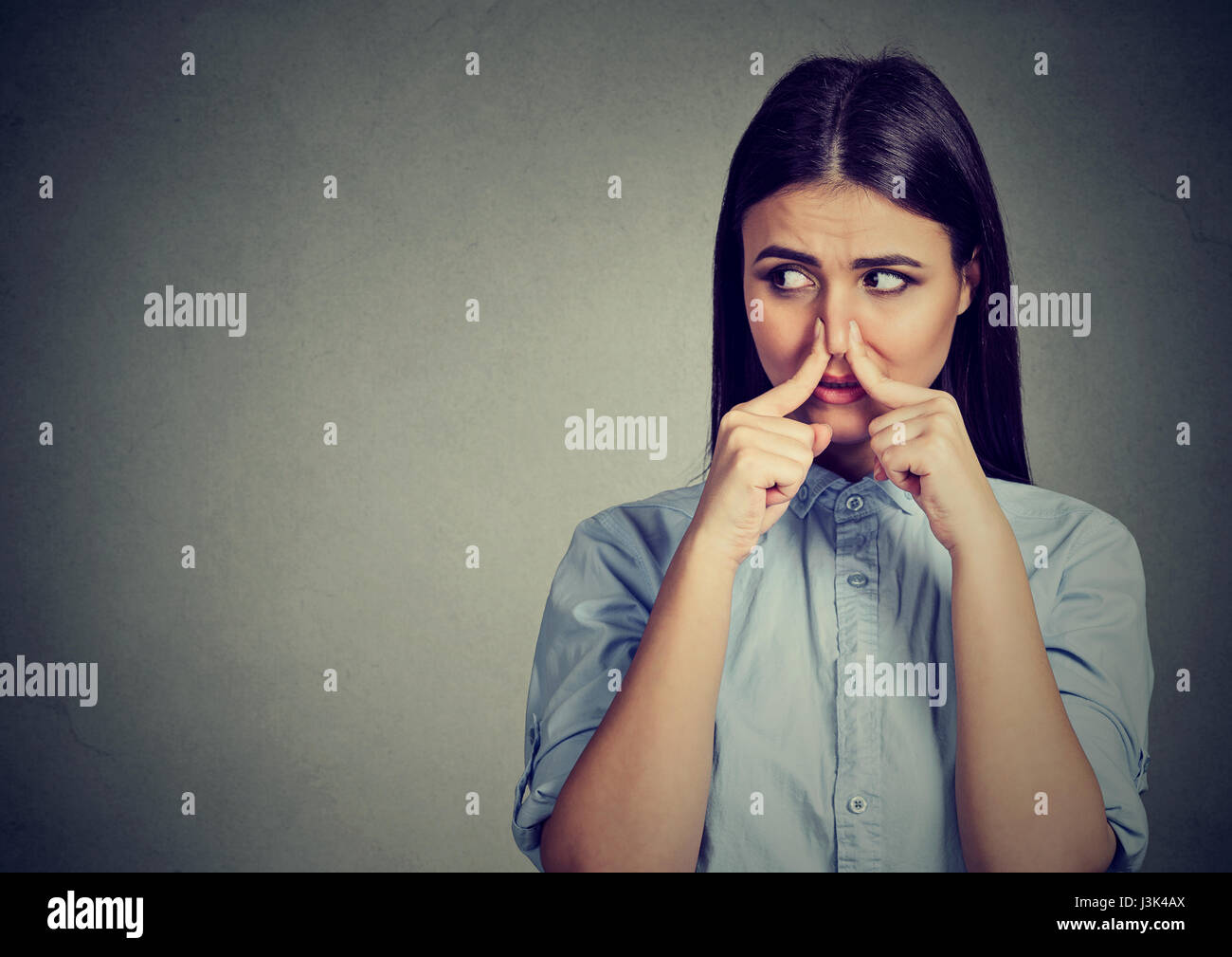 Woman pinches nose with fingers looks with disgust away something stinks bad smell isolated on gray background. Stock Photo