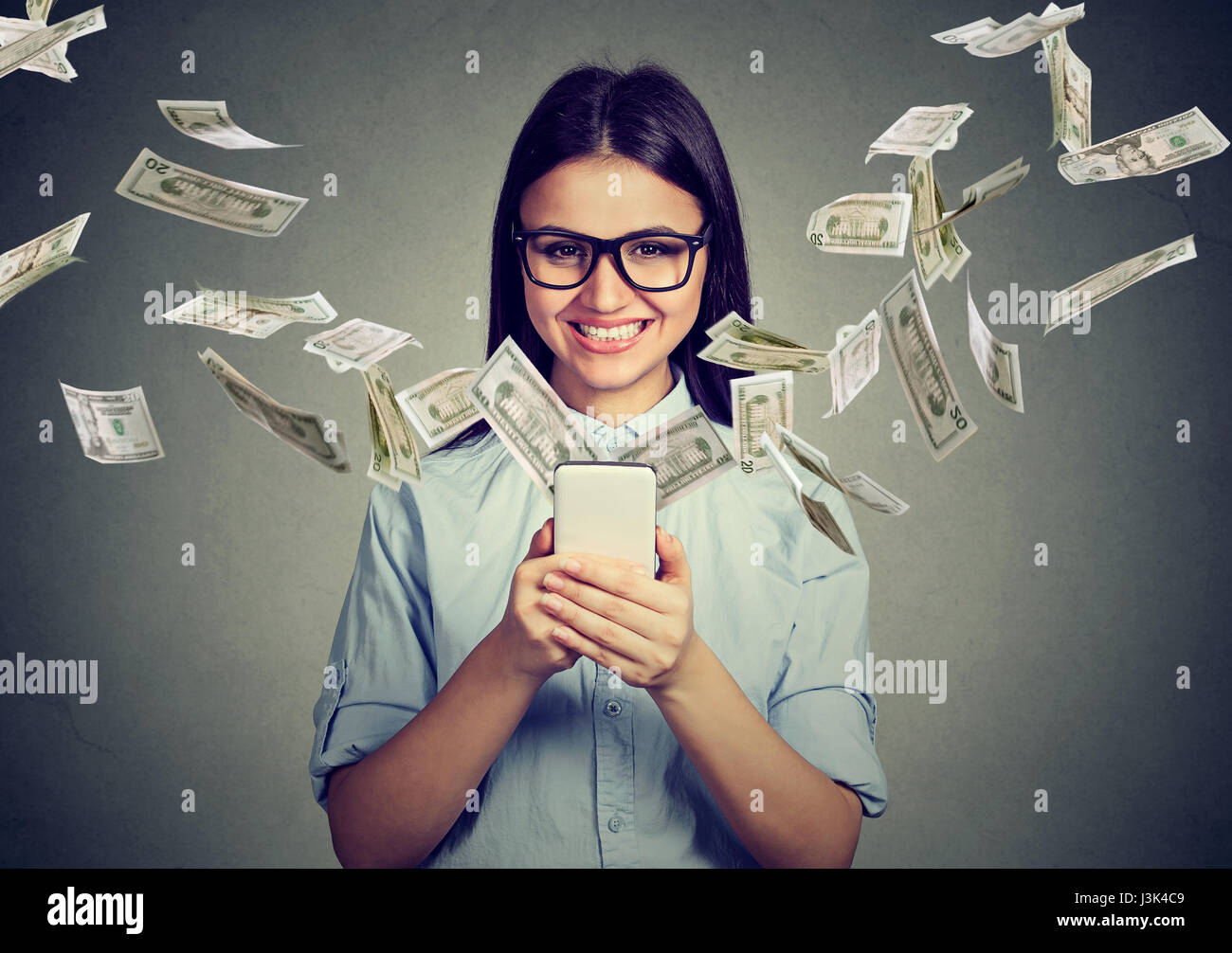 Online banking money transfer, e-commerce concept. Happy young woman in glasses using smartphone with dollar bills flying away from screen isolated on Stock Photo