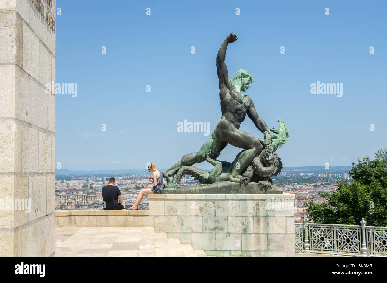 Two people looking at their smartphone s instead of the view from Gellert Hill in Budapest while sitting next to a statue of a man slaying a dragon. Stock Photo