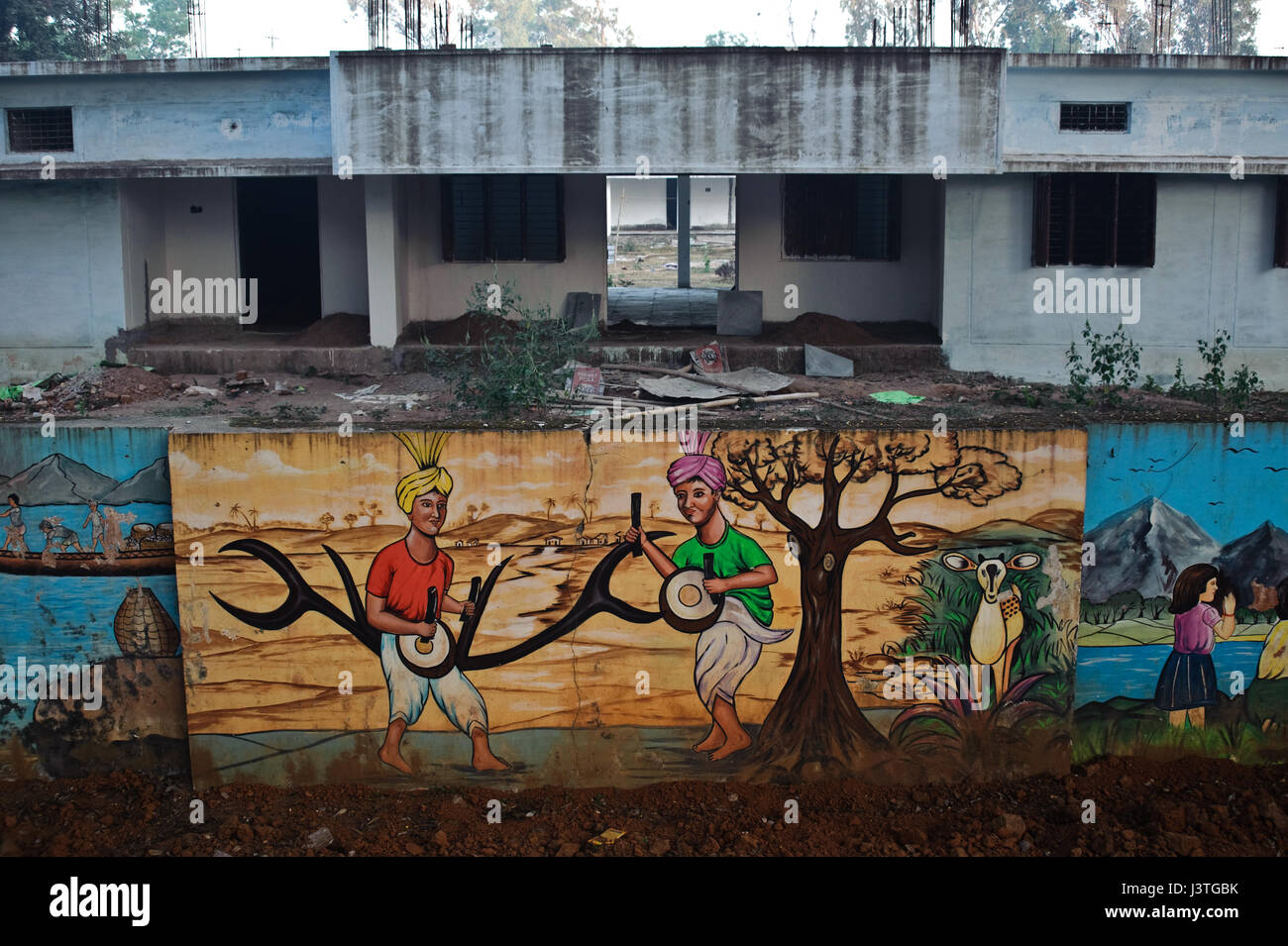 Mural painting depicting tribesmen playing drums in the nature. In the background, school under repair ( India) Stock Photo