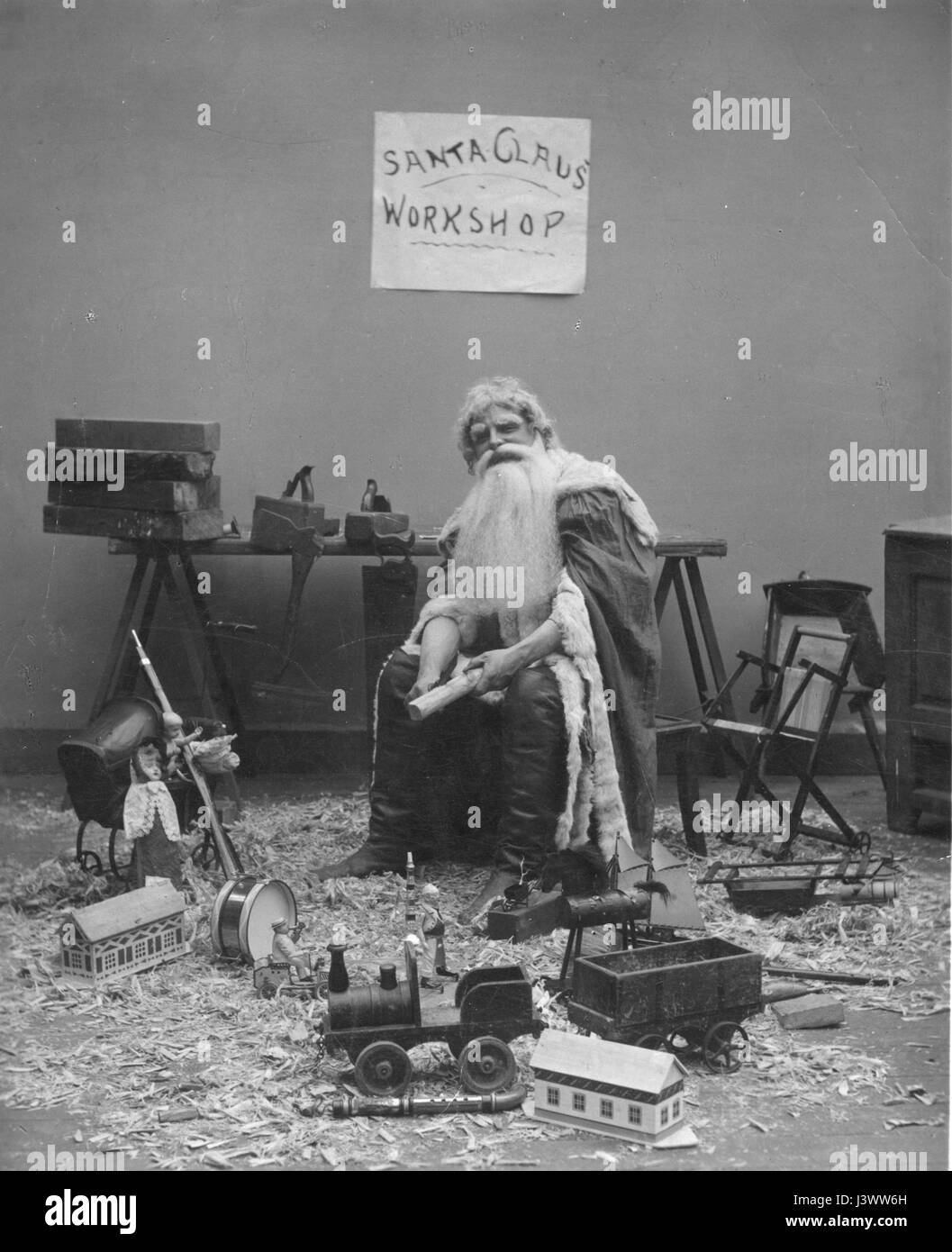 'Santa Claus' Workshop'  Santa is whittling a new toy, while sitting in his workshop, c. 1908.  Completed toys around him are a drum, simple train engine with car, dolls and a toy baby carriage, a small hobby horse, a two-hand whistle, other small toys and lots of wood shavings. His work table holds wood planes, wood to be carved, and an ax and a saw hang from the table's side.  To see my other Christmas-related vintage images, Search:  Prestor  vintage  holiday Stock Photo