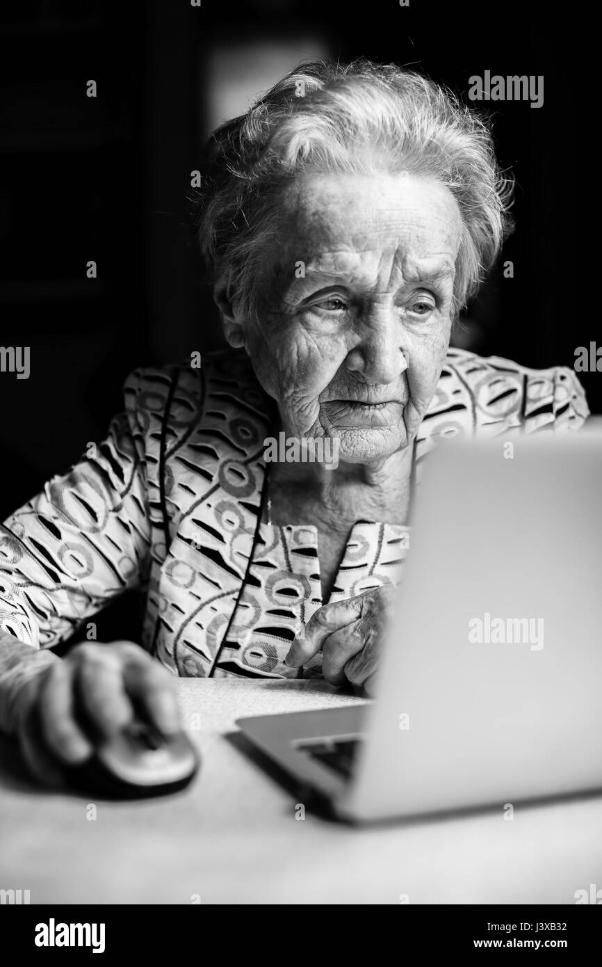 An elderly woman sitting at a table with a laptop, black-and-white photo. Stock Photo