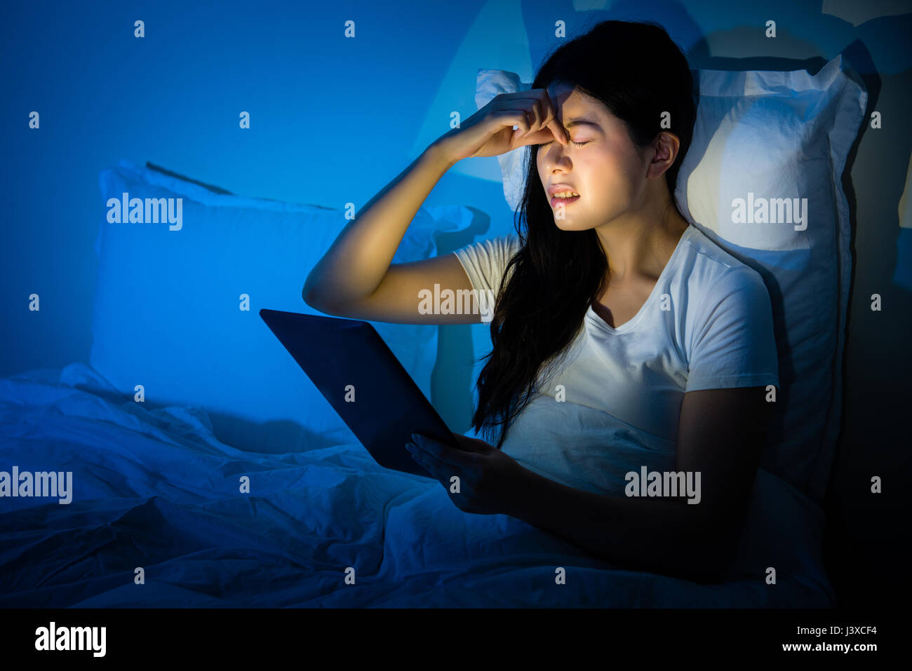 Asian woman feeling eye pain high intraocular pressure when using tablet at night sitting on bed. mixed race asian chinese model. Stock Photo