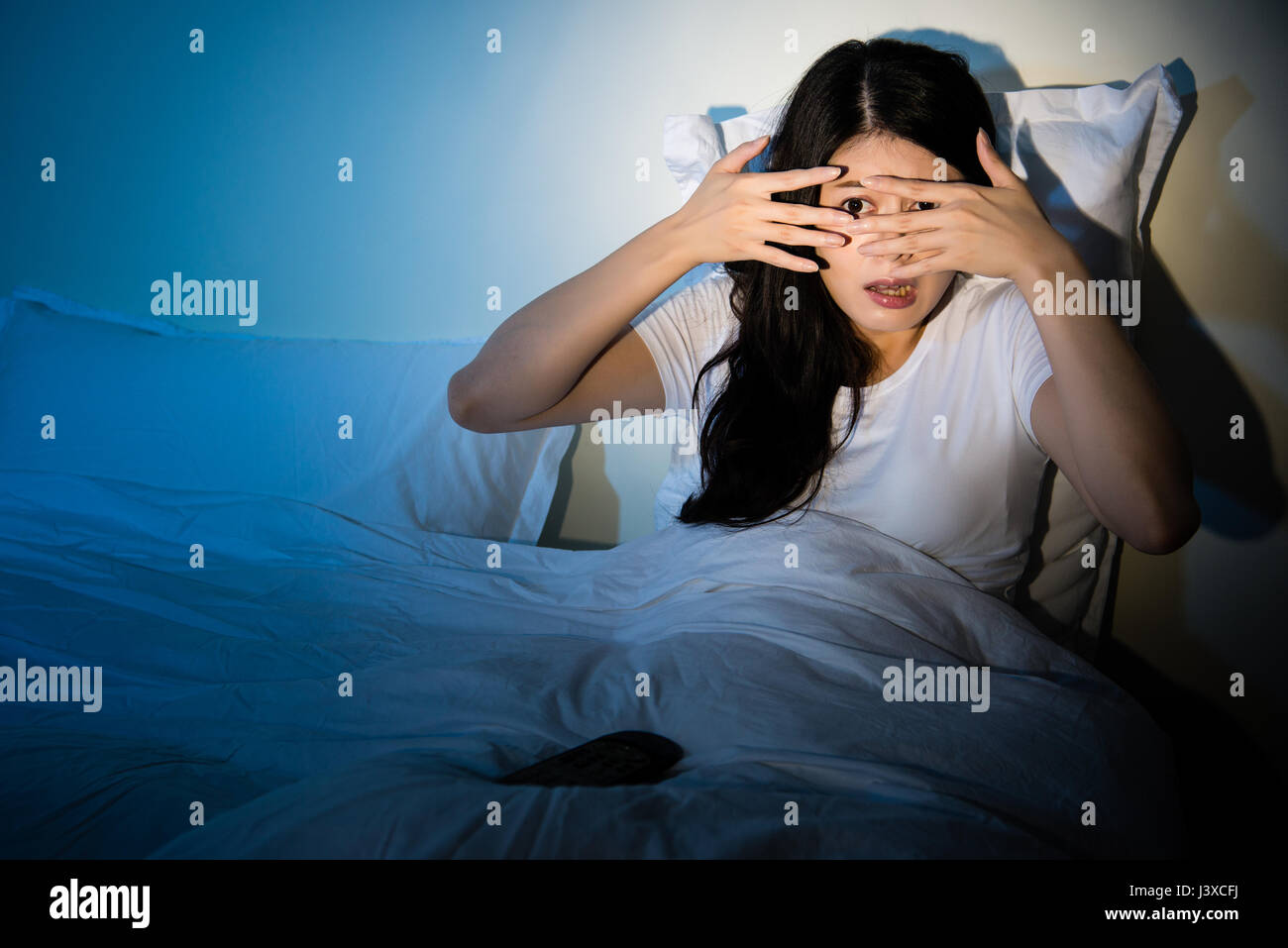 young girl sitting on a bed watching a horror movie scary to cove her eyes on TV. mixed race asian chinese model. Stock Photo