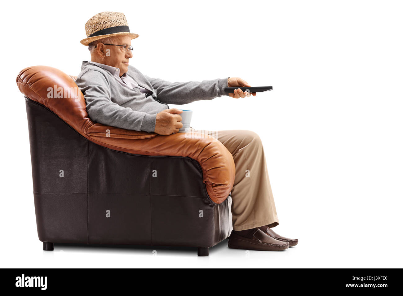 Bored elderly man holding a cup and a remote sitting in an armchair and watching television isolated on white background Stock Photo