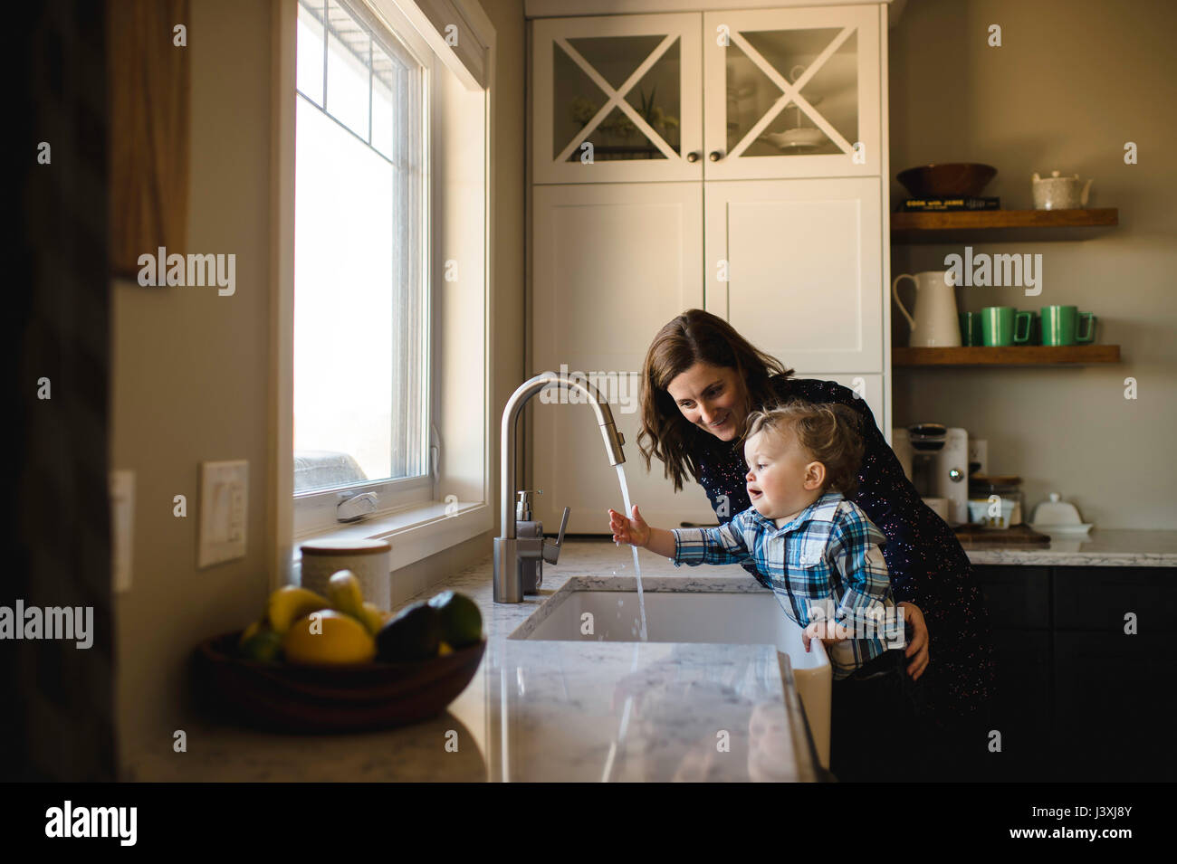 Mother helping toddler son wash hands at kitchen sink Stock Photo