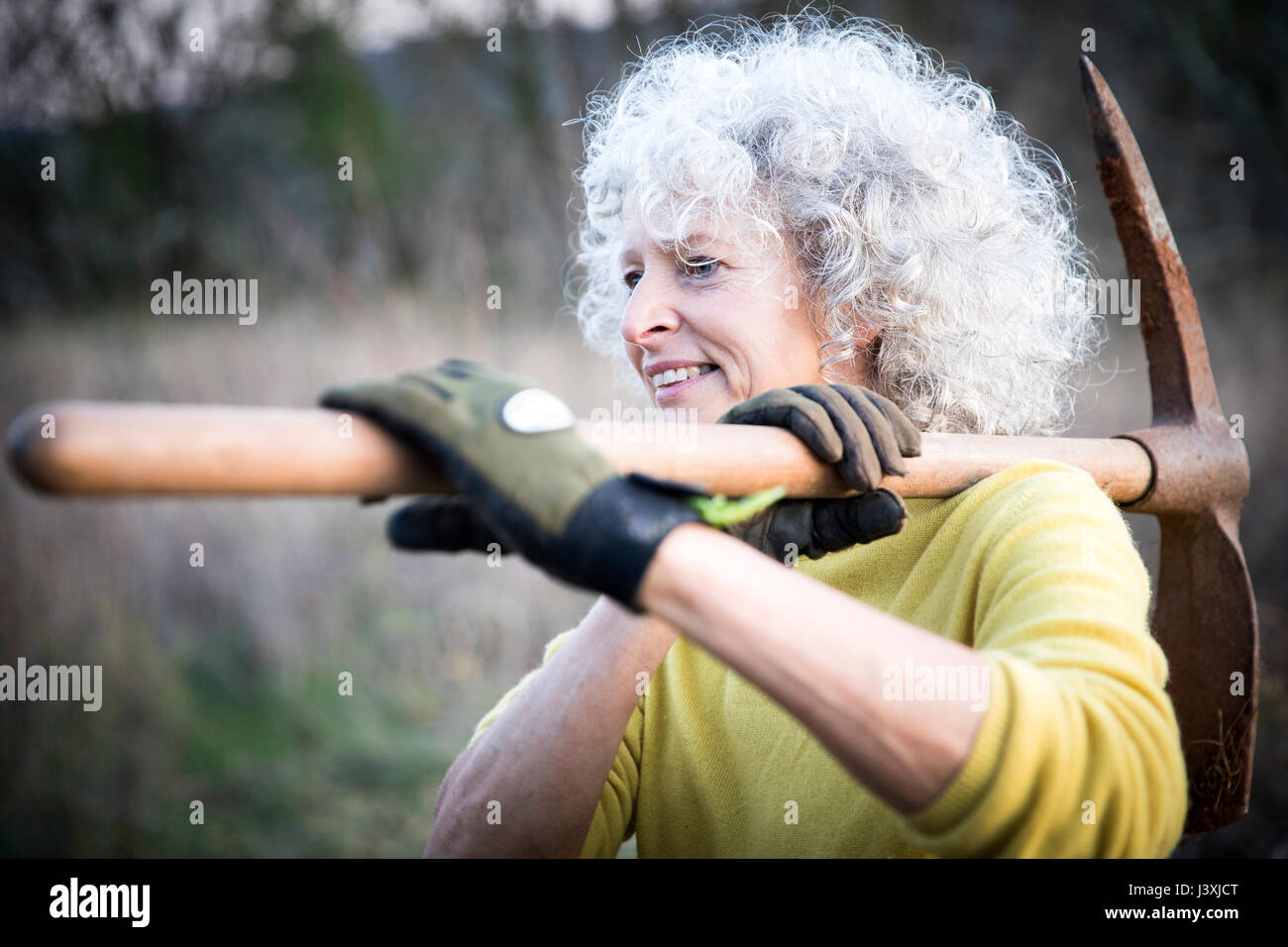 Mature woman with grey hair carrying pick axe over her shoulder in woodland Stock Photo