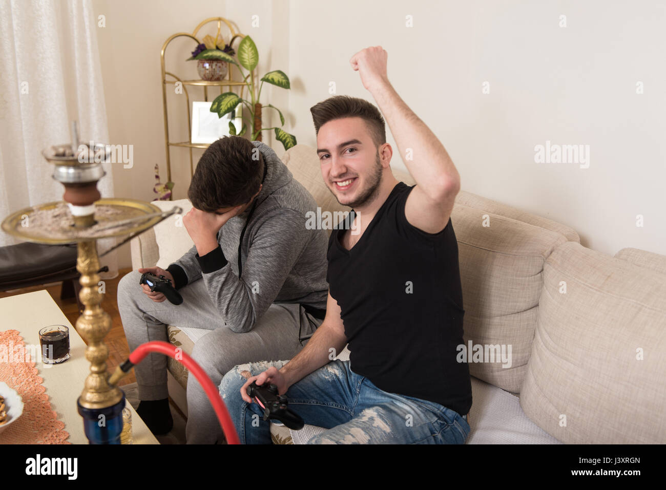 Two Young Gamers Sitting Together On Sofa And Playing Video Games At
