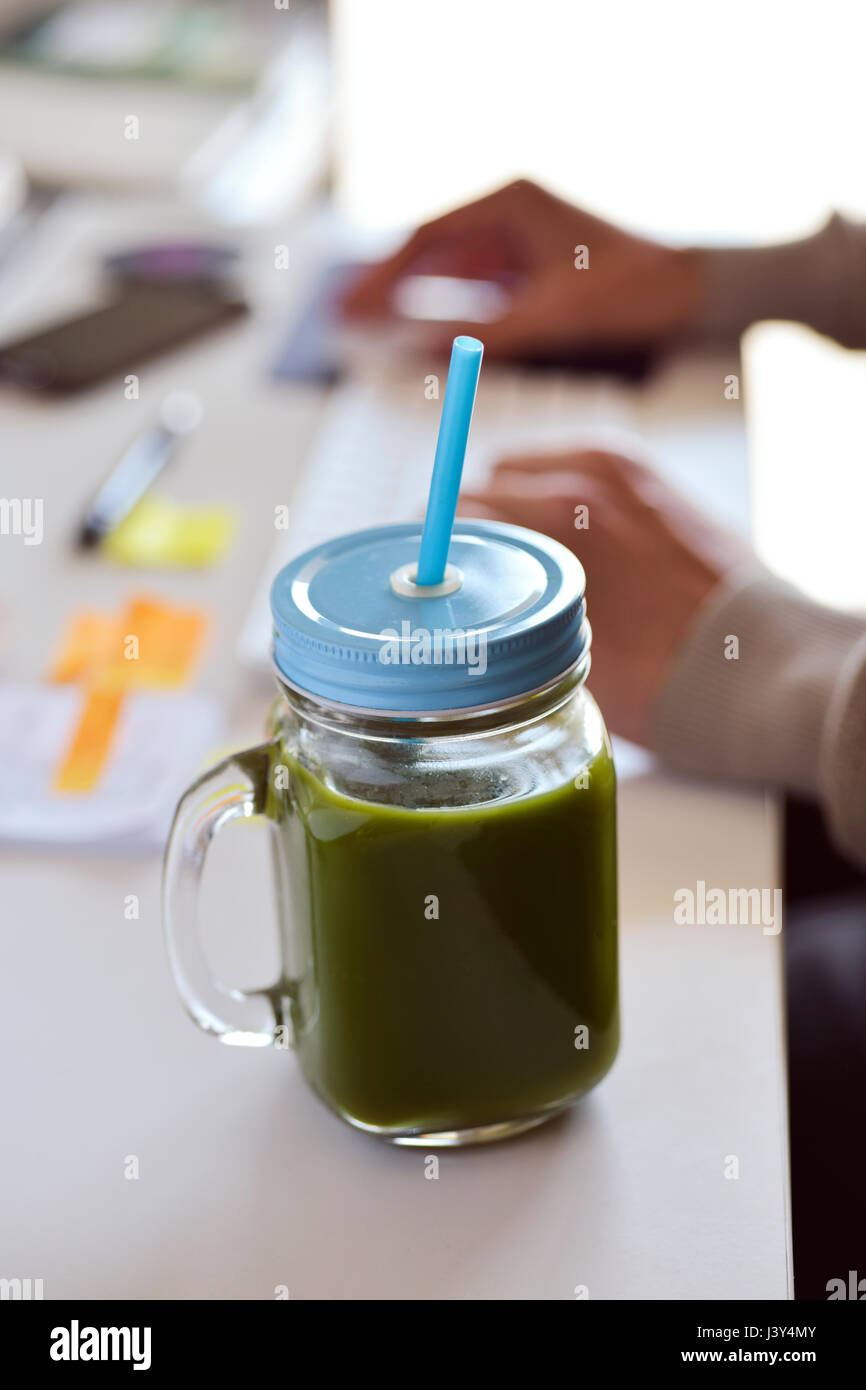 closeup of a green detox smoothie served in a glass jar on an office desk, and a young caucasian man using his computer in the background Stock Photo