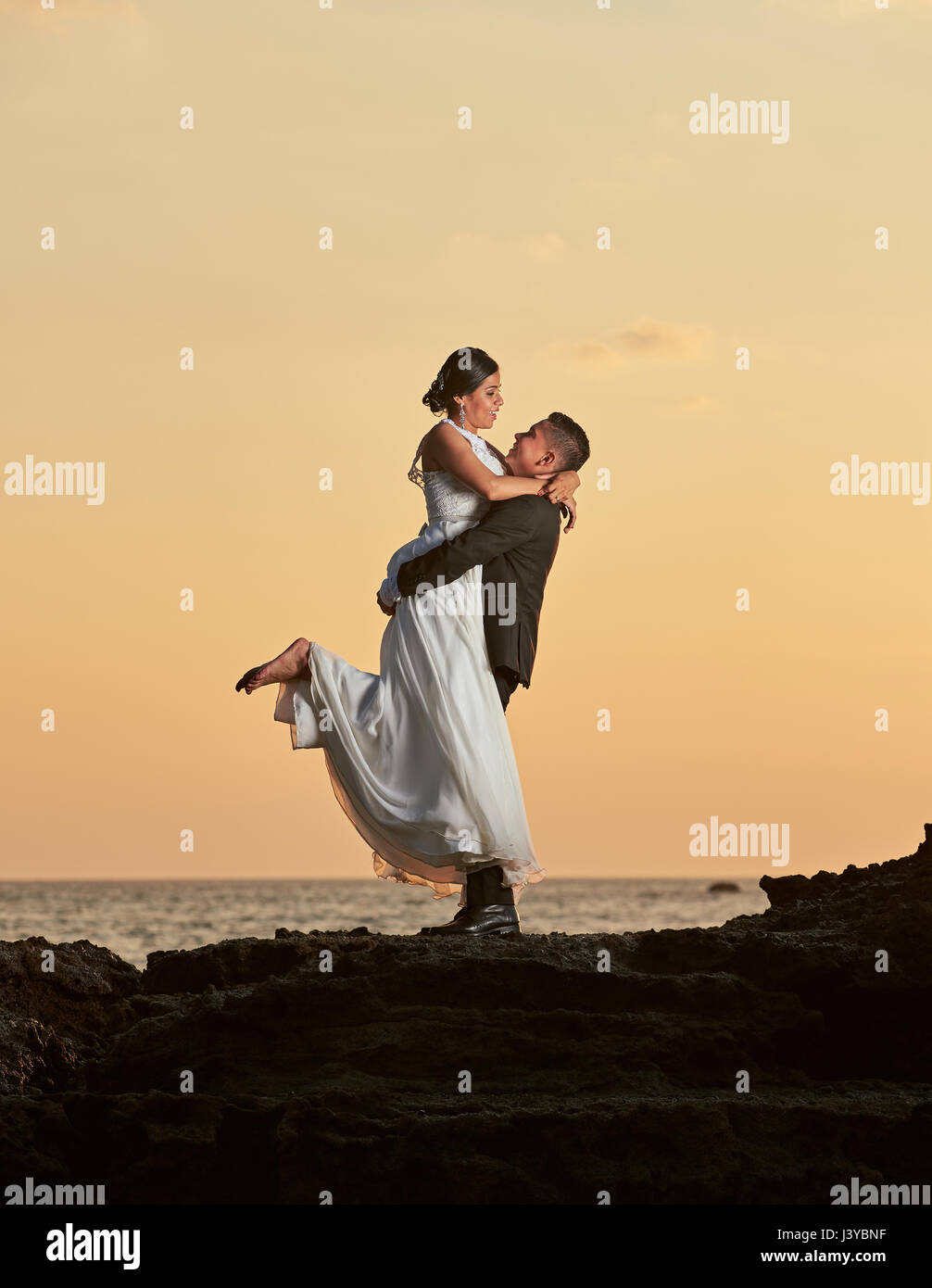 Groom lifting bride on orange sunset background Stock Photo