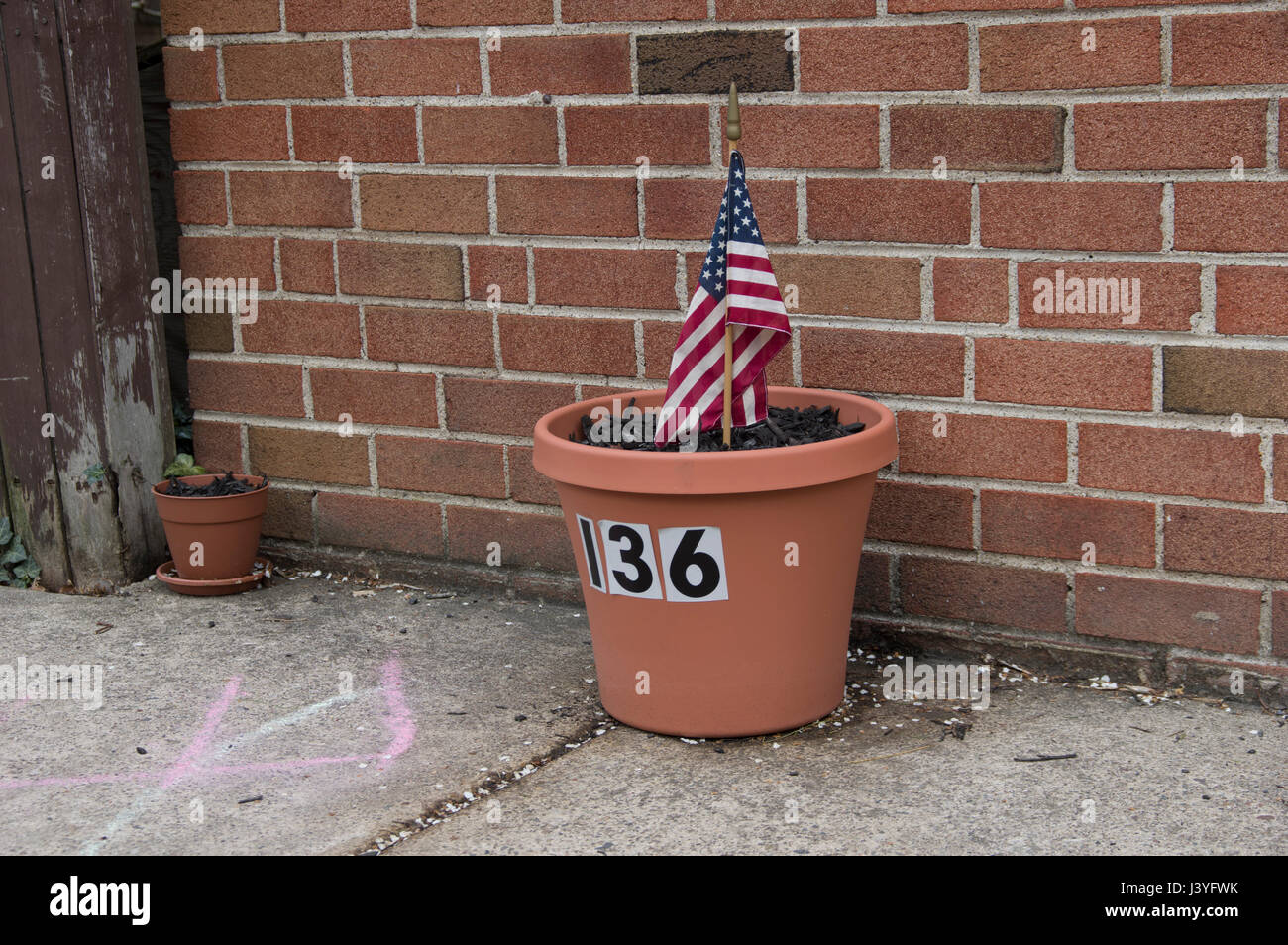 flower pot against a brick wall with the number 136 on it and an small American flag stuck in it Stock Photo