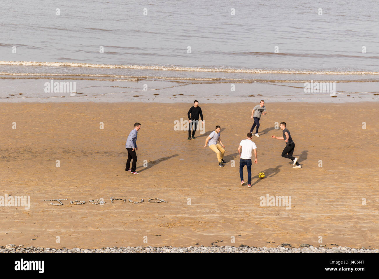 Six young men playing football soccer on the beach including a name made out of beach pebbles Norfolk UK Stock Photo