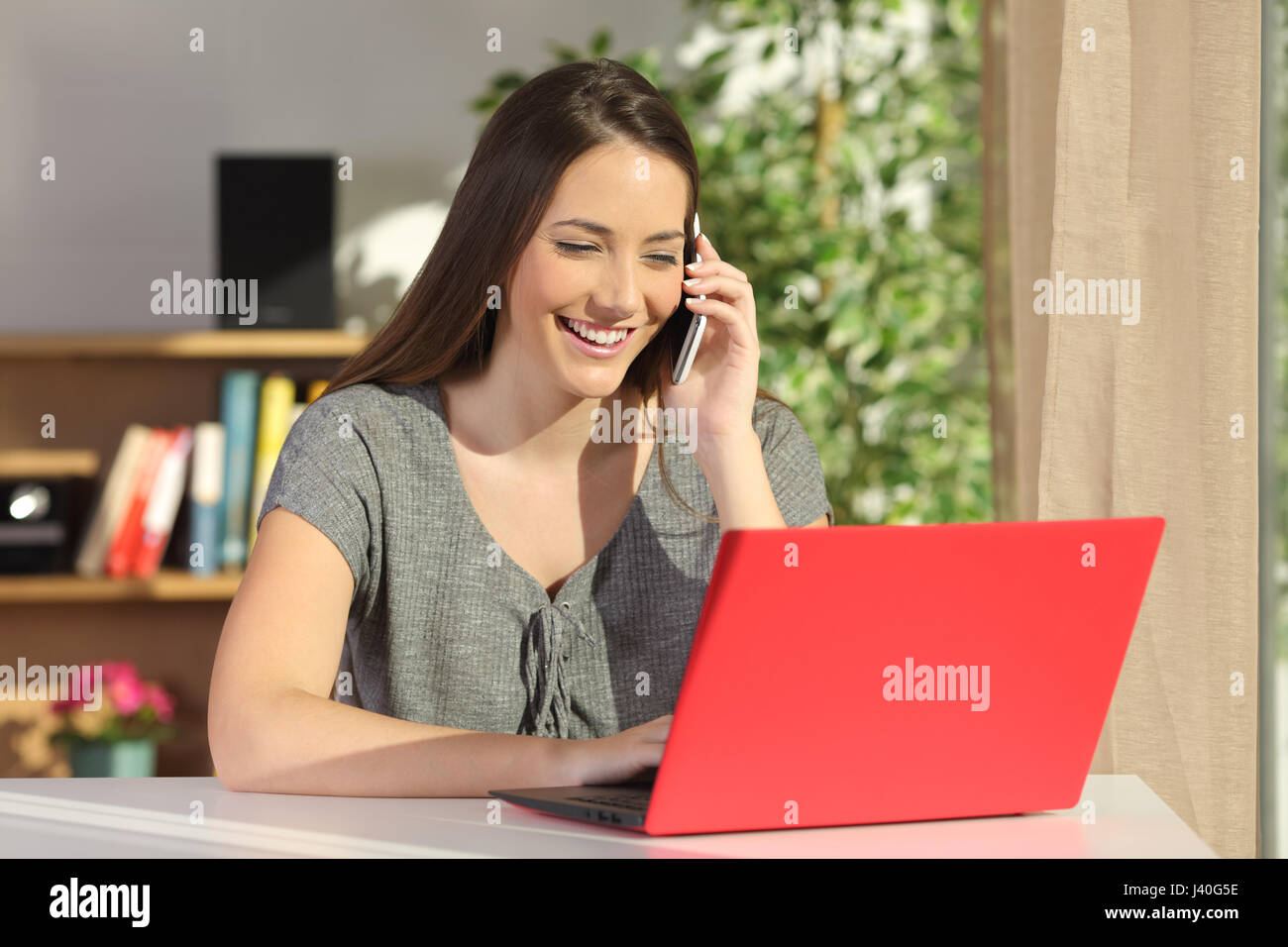 Attractive housewife using a red laptop and calling to the assistance service on line with a mobile phone in a table in a house interior Stock Photo