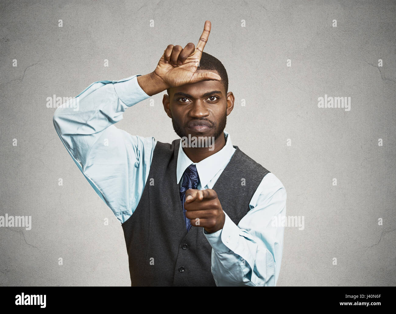 Closeup portrait angry mad young Unhappy Man, student displaying Loser Sign on forehead, pointing at you with disgust, isolated grey background. Negat Stock Photo