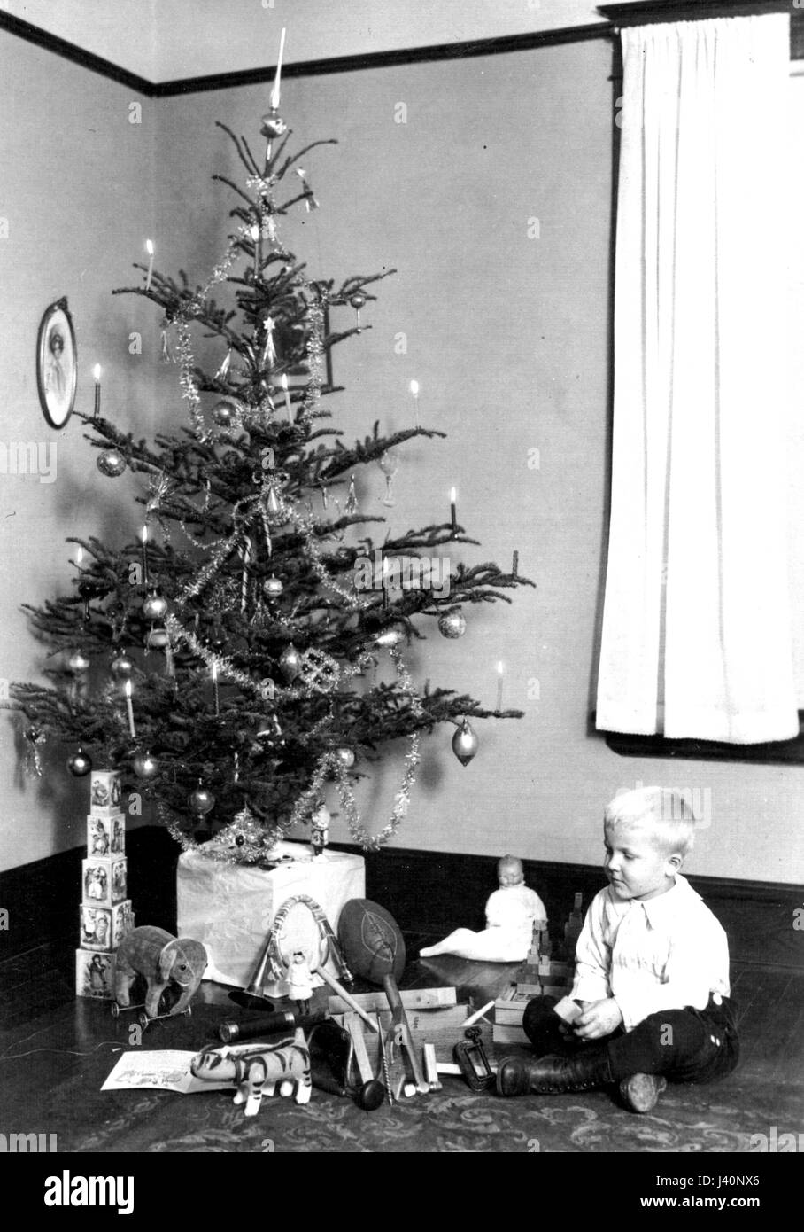 A young boy seated next to the family Christmas tree, which has lighted candles, c.1915.  Many gifts are laid out, including a toy elephant, toy tools, a football, children's blocks, toy horn, a doll, etc.   To see my other Christmas-related vintage images, Search:  Prestor  vintage  holiday Stock Photo