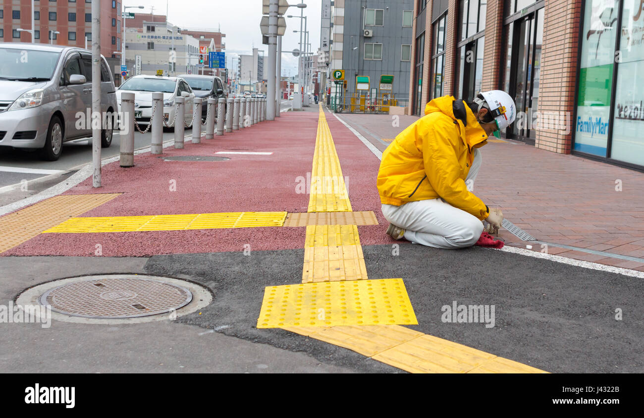Hakodate, Japan - April 3th, 2017: A Japanese worker wearing yellow safety workwear, is on knees working on the sidewalk across the JR Hakodate Train  Stock Photo