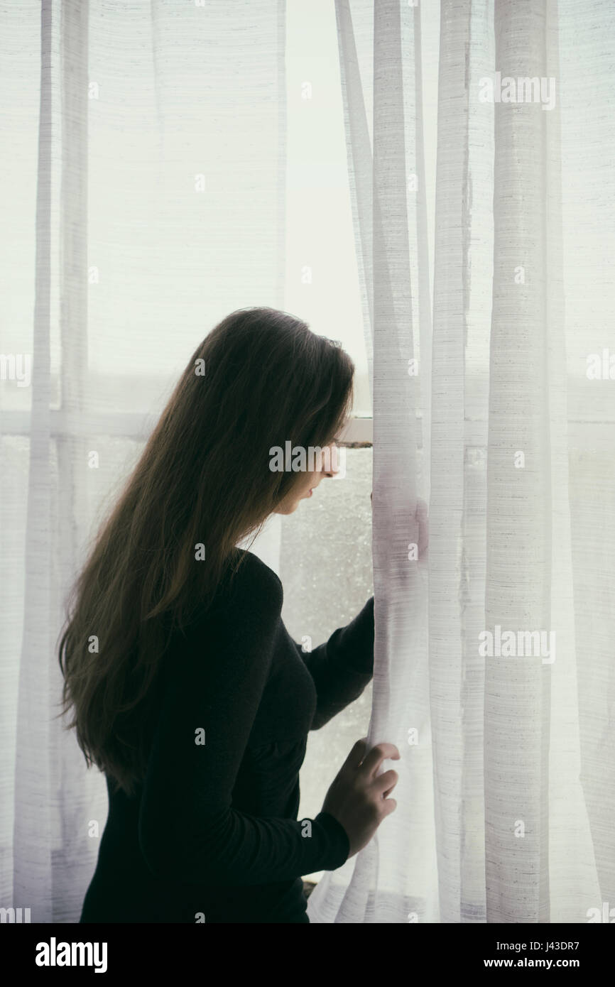 Resr view of a woman moving curtains to look out of the window waiting