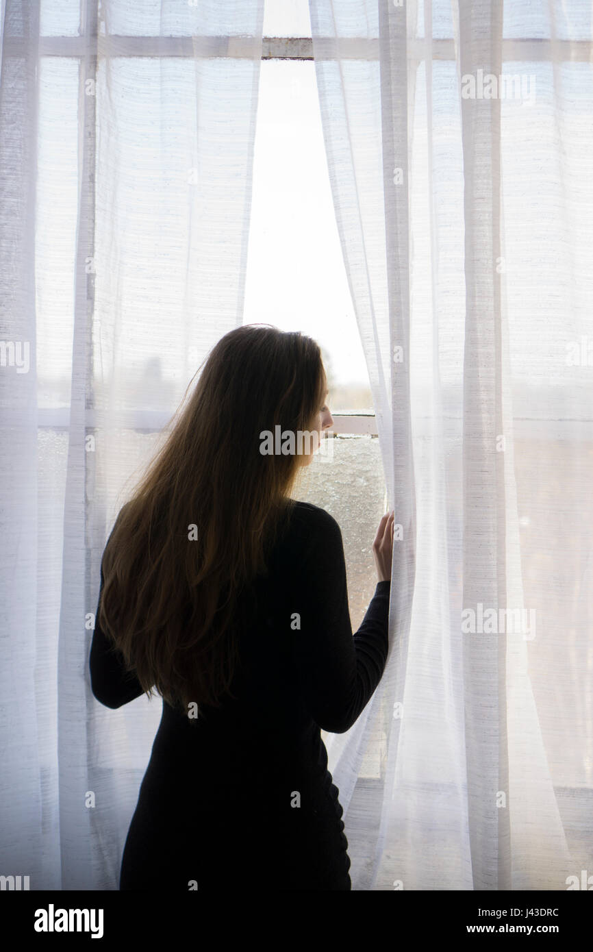 Resr view of a woman moving curtains to look out of the window waiting